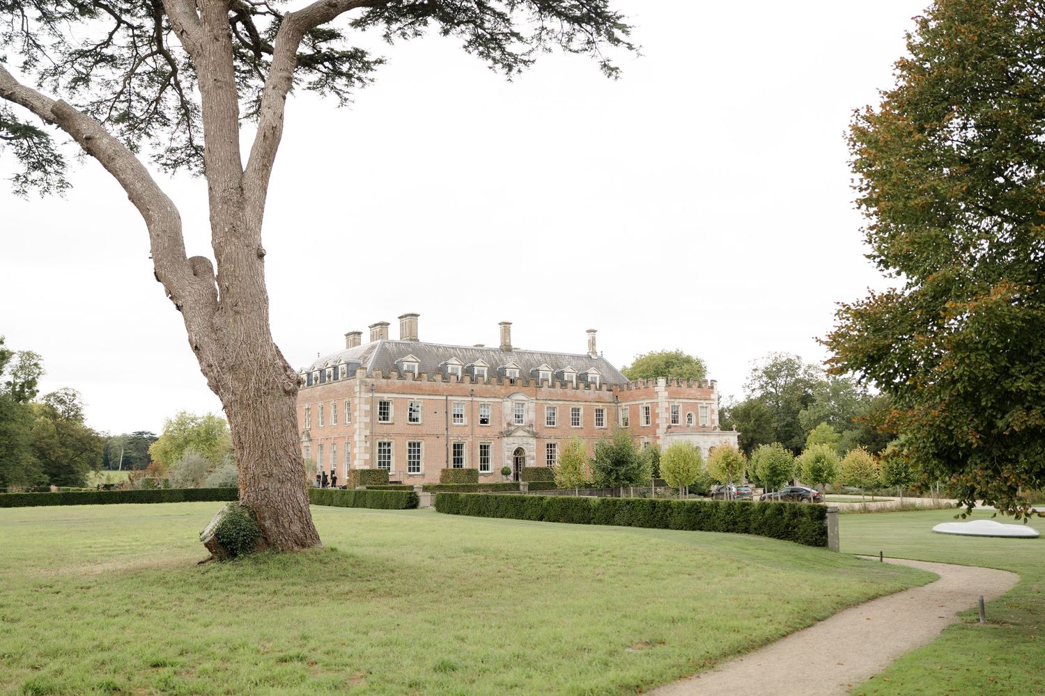 Grand French-style country manor house with manicured hedges, sweeping lawns, and a large tree in the foreground.