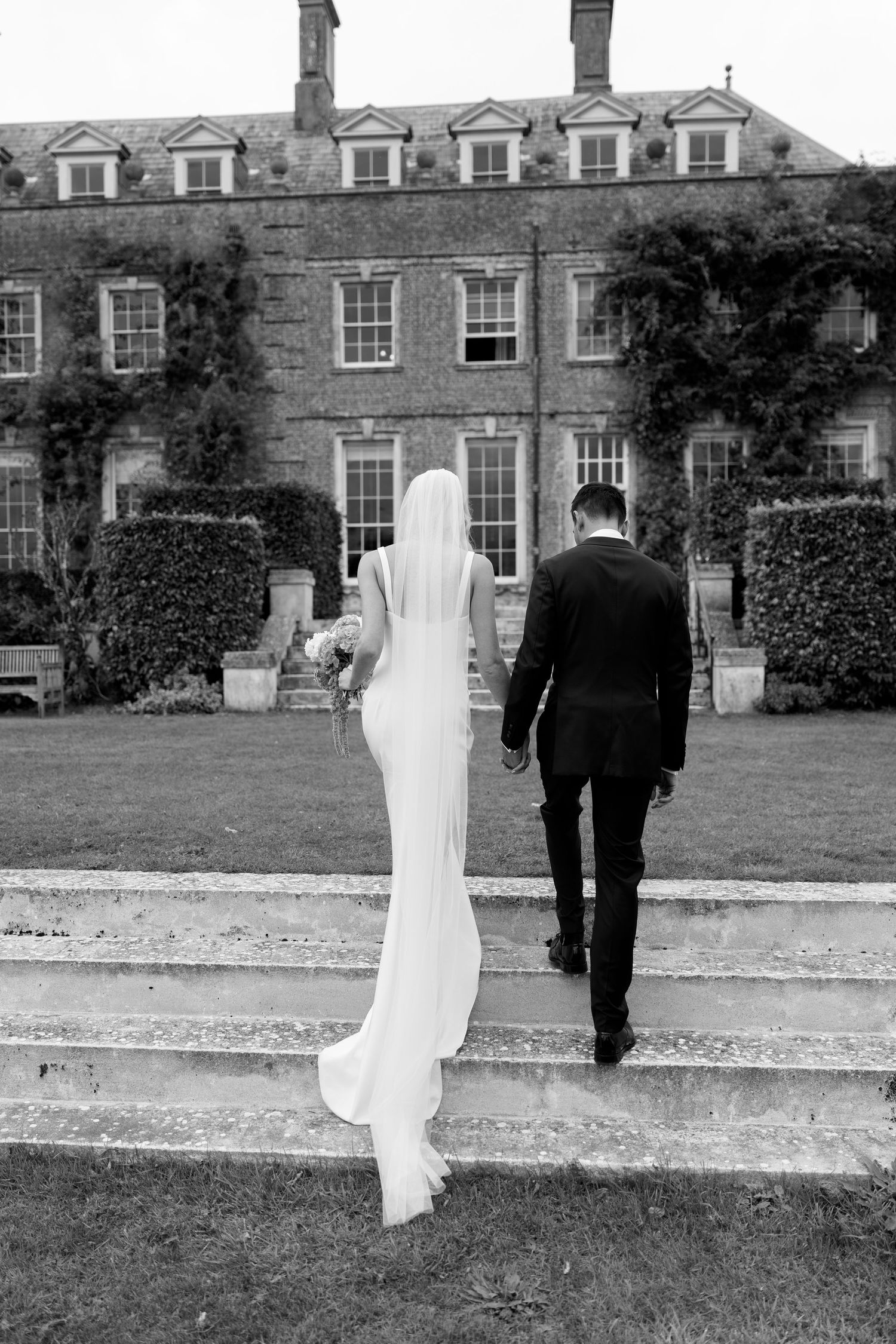 Black and white photo of newlyweds walking toward ivy-covered English country house estate.