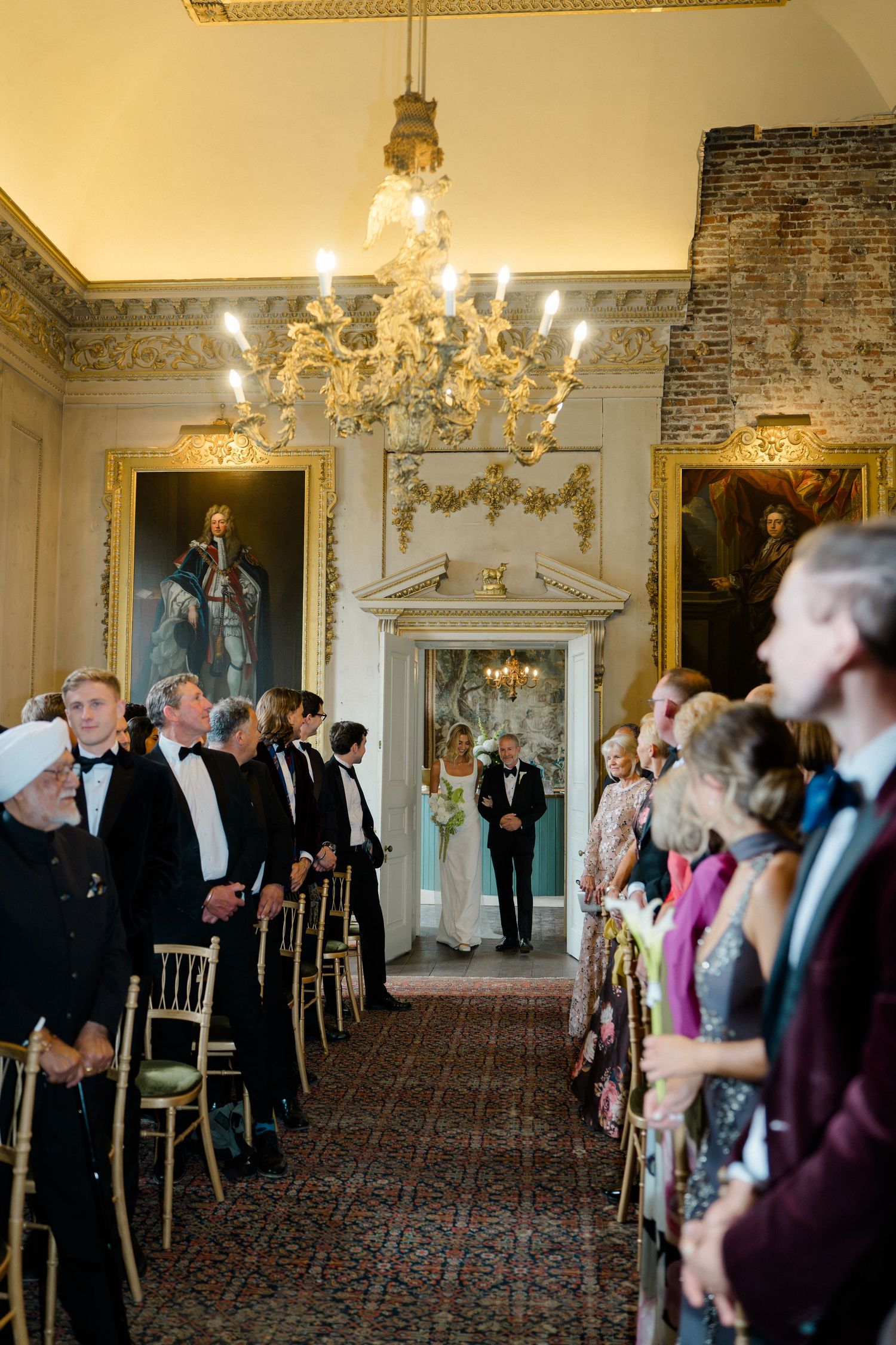 Bride walking down the aisle in elegant ceremony room with chandeliers and portrait paintings.
