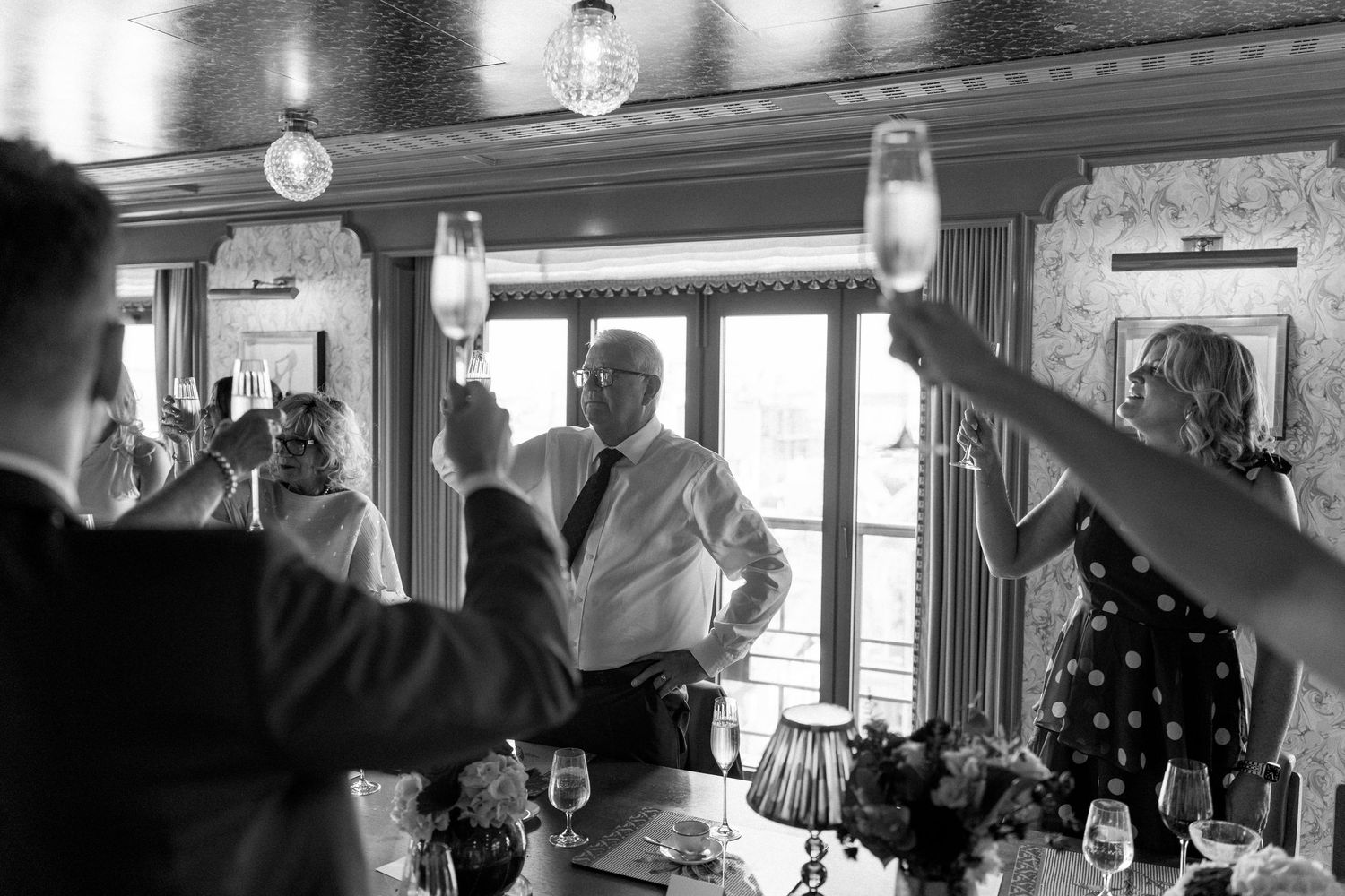 Black and white photo of wedding guests raising champagne glasses in a toast at reception dinner.