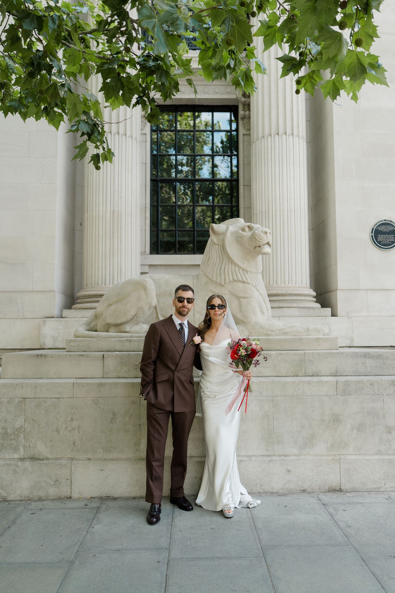 Bride and groom pose in sunglasses outside a classical building with lion sculpture and columns on wedding day.
