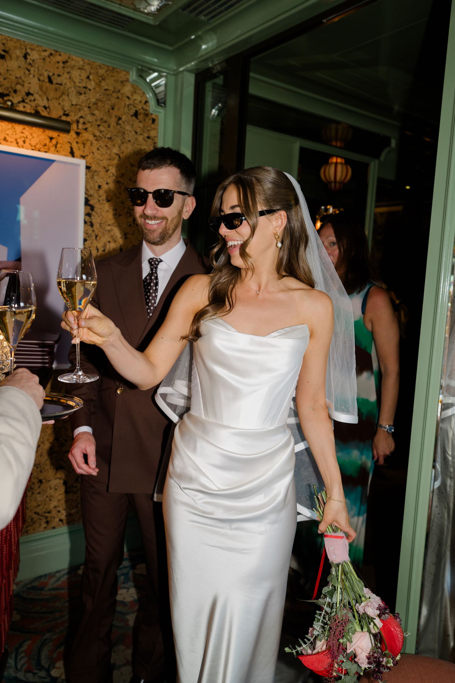 Bride and groom in sunglasses toasting champagne in white satin wedding dress and brown suit.