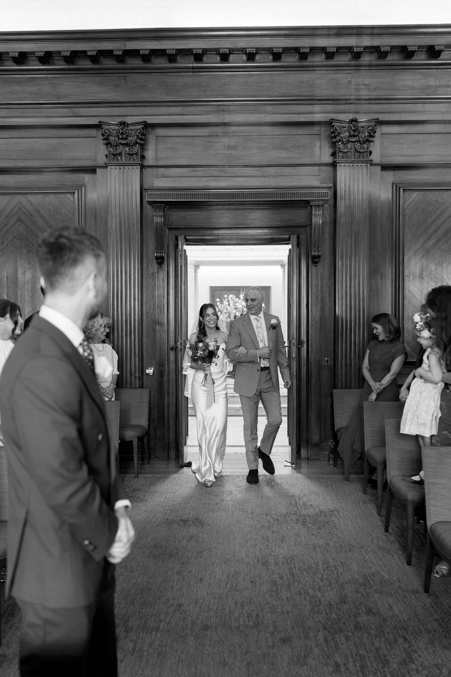 Bride walks down the aisle with escort in elegant wood-paneled ceremony hall, guests standing on both sides.