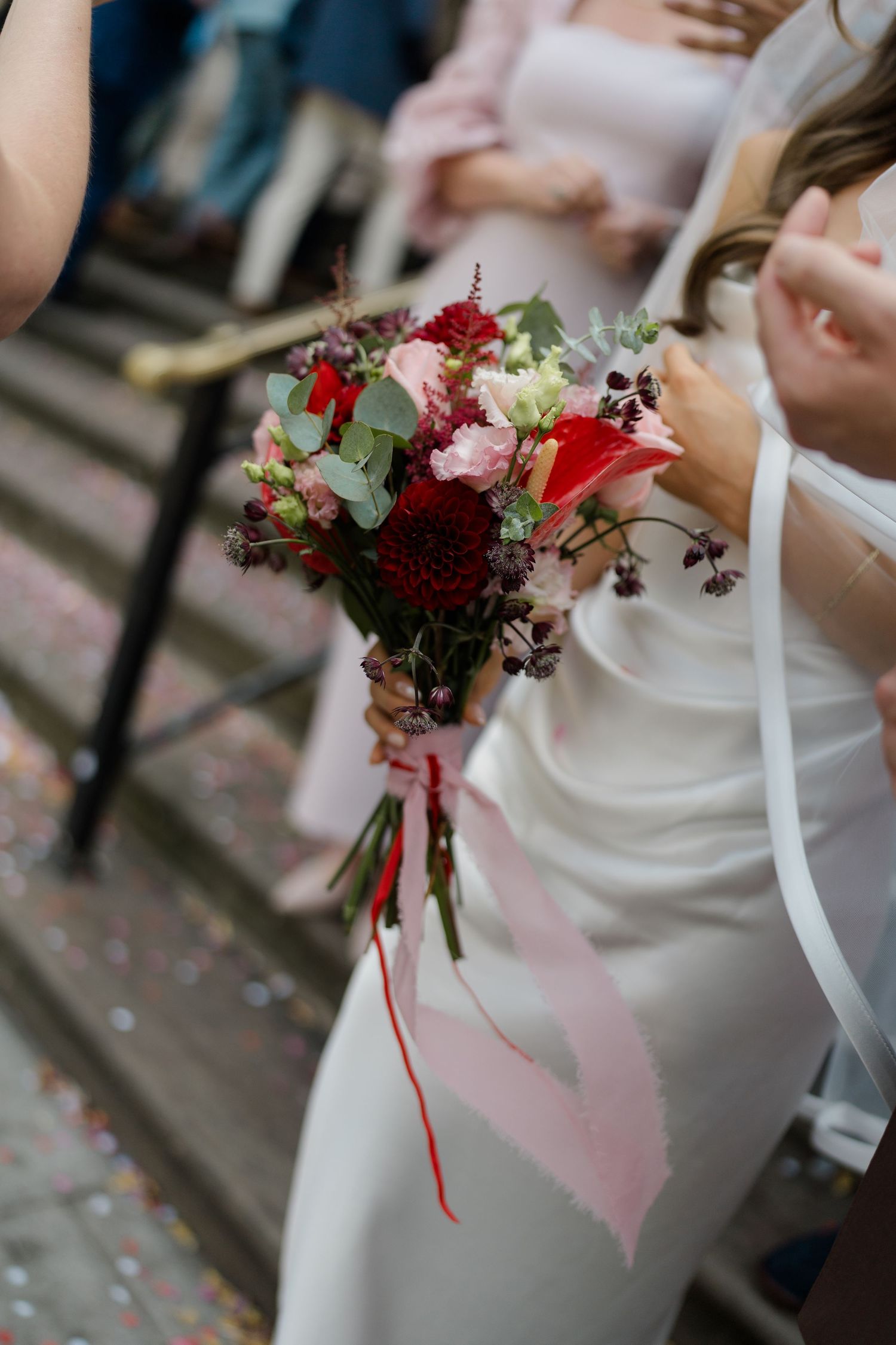Bride holds vibrant red and pink wedding bouquet with ribbon on stone steps with confetti scattered around.