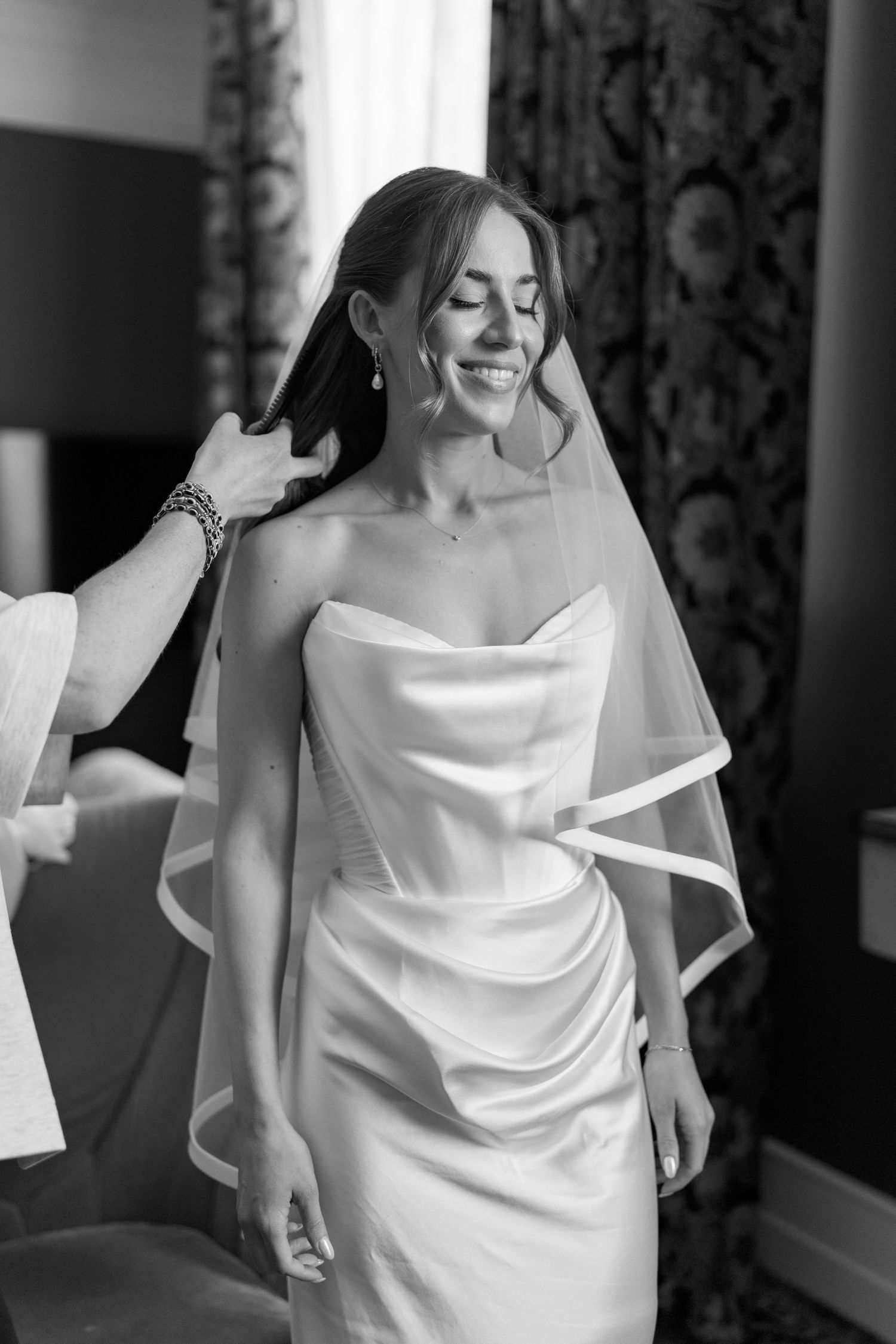 Smiling bride in strapless satin gown and veil getting hair styled in black and white wedding preparation photo.