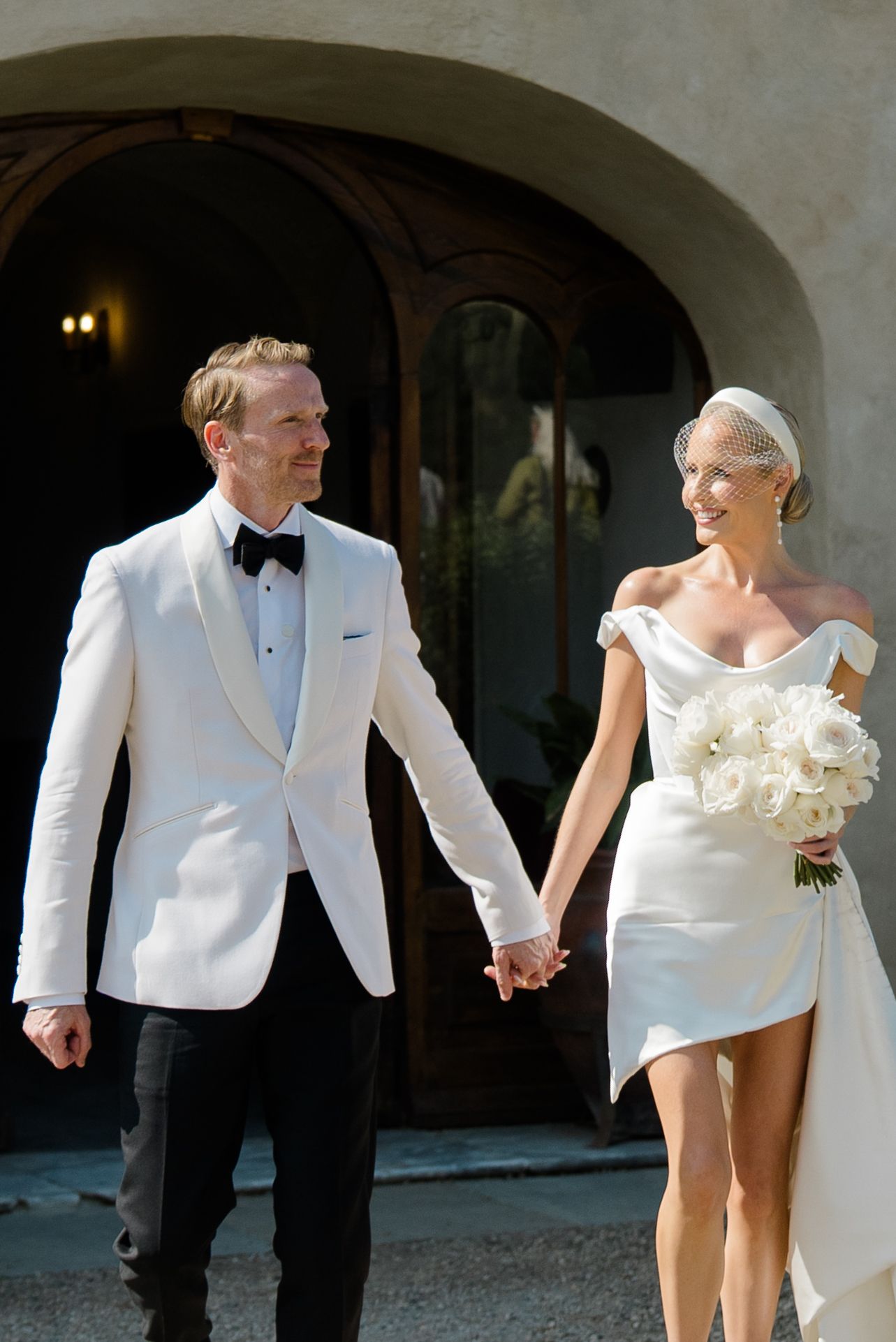 Smiling bride and groom in white tuxedo hold hands walking under stone archway on wedding day.