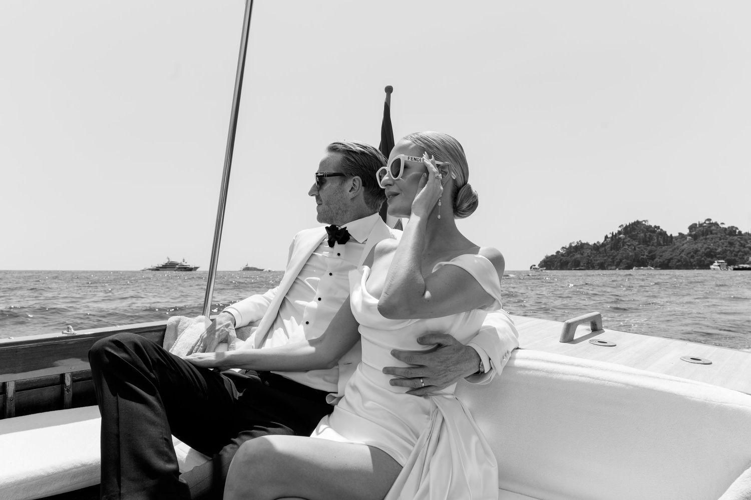 Newlyweds embrace on boat deck in black and white photo with calm ocean in background.