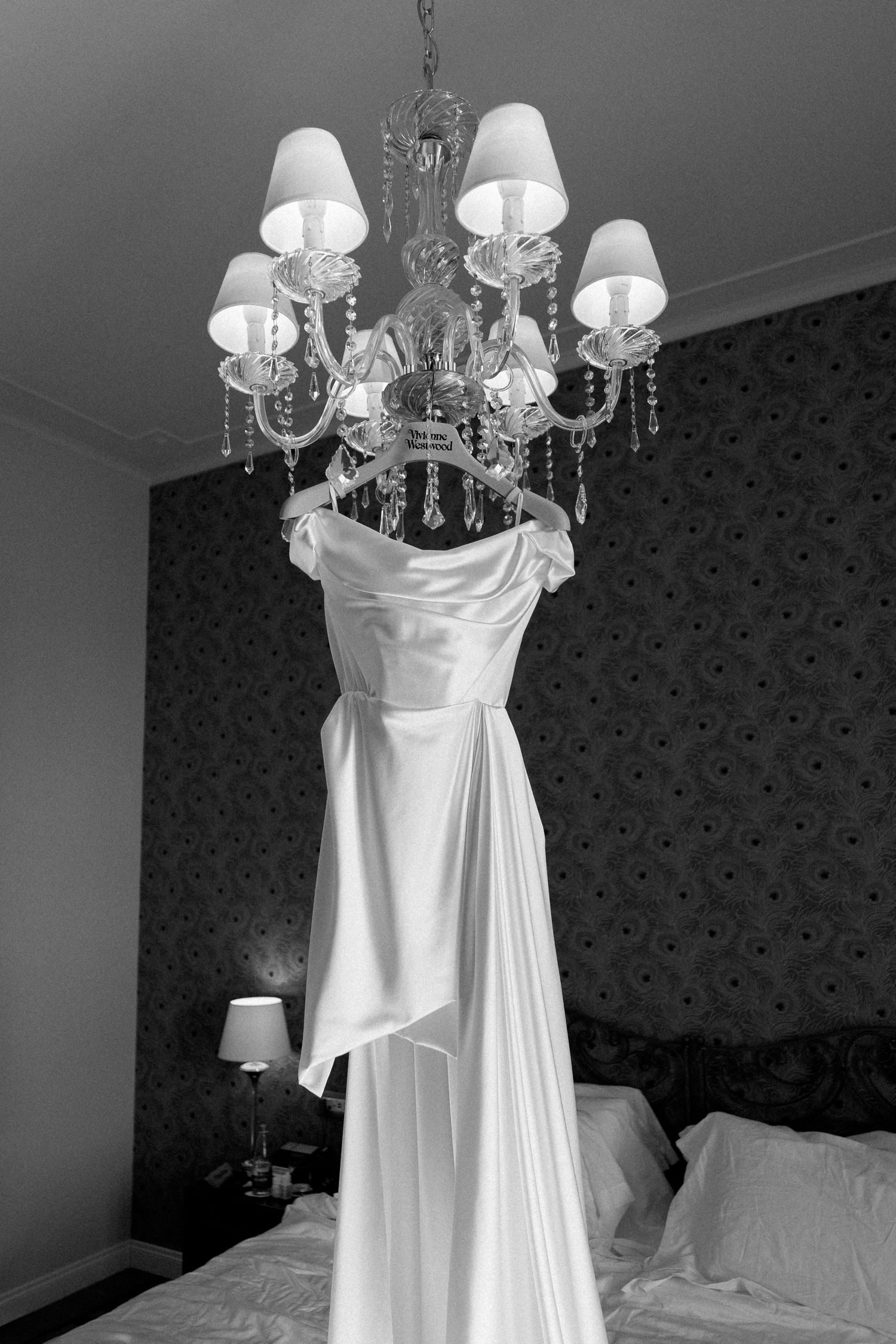 Black and white photo of an elegant white wedding dress hanging from a crystal chandelier in a luxury bedroom.