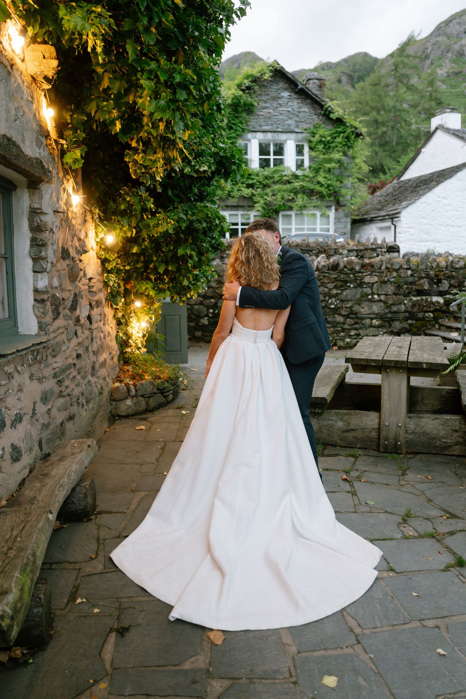 Couple dances on stone patio at dusk with string lights and mountain backdrop.