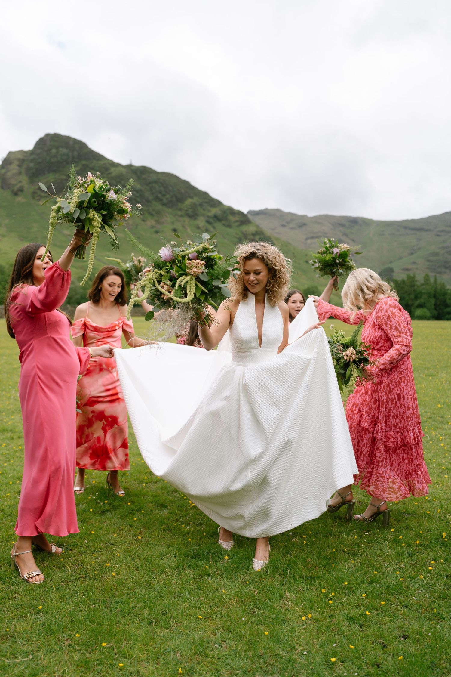 Group in coral dresses hold up a flowing white wedding dress in a scenic mountain meadow.