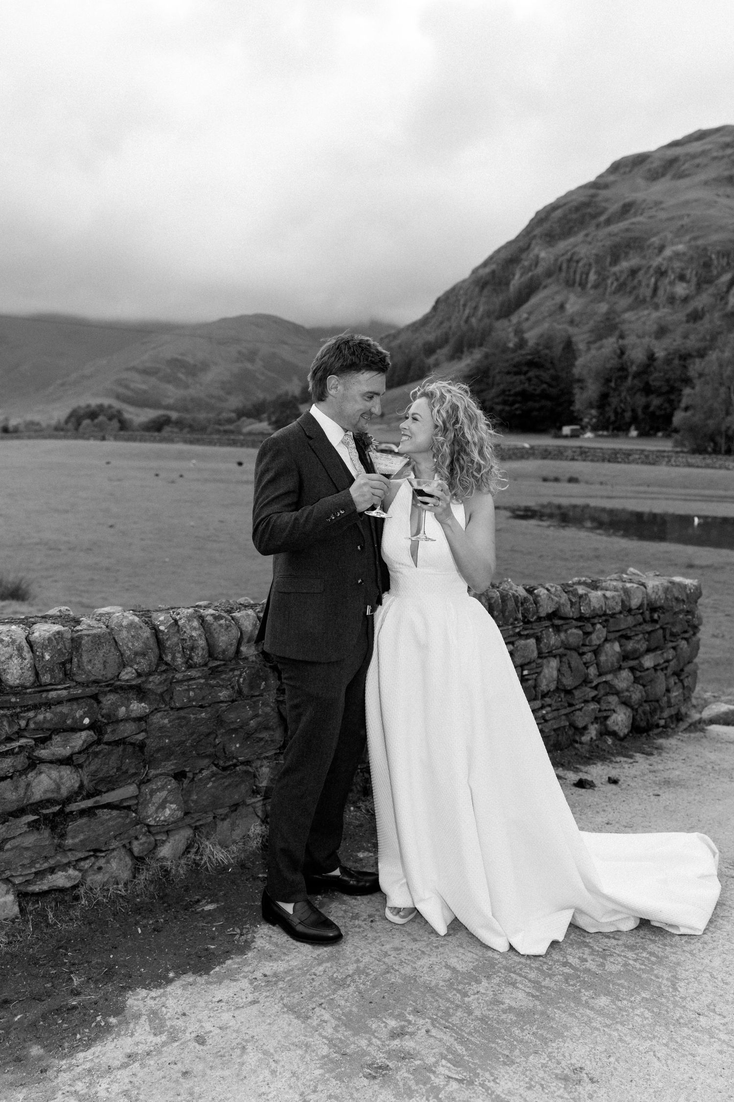 Couple shares intimate moment by stone wall with scenic mountain backdrop in black and white.
