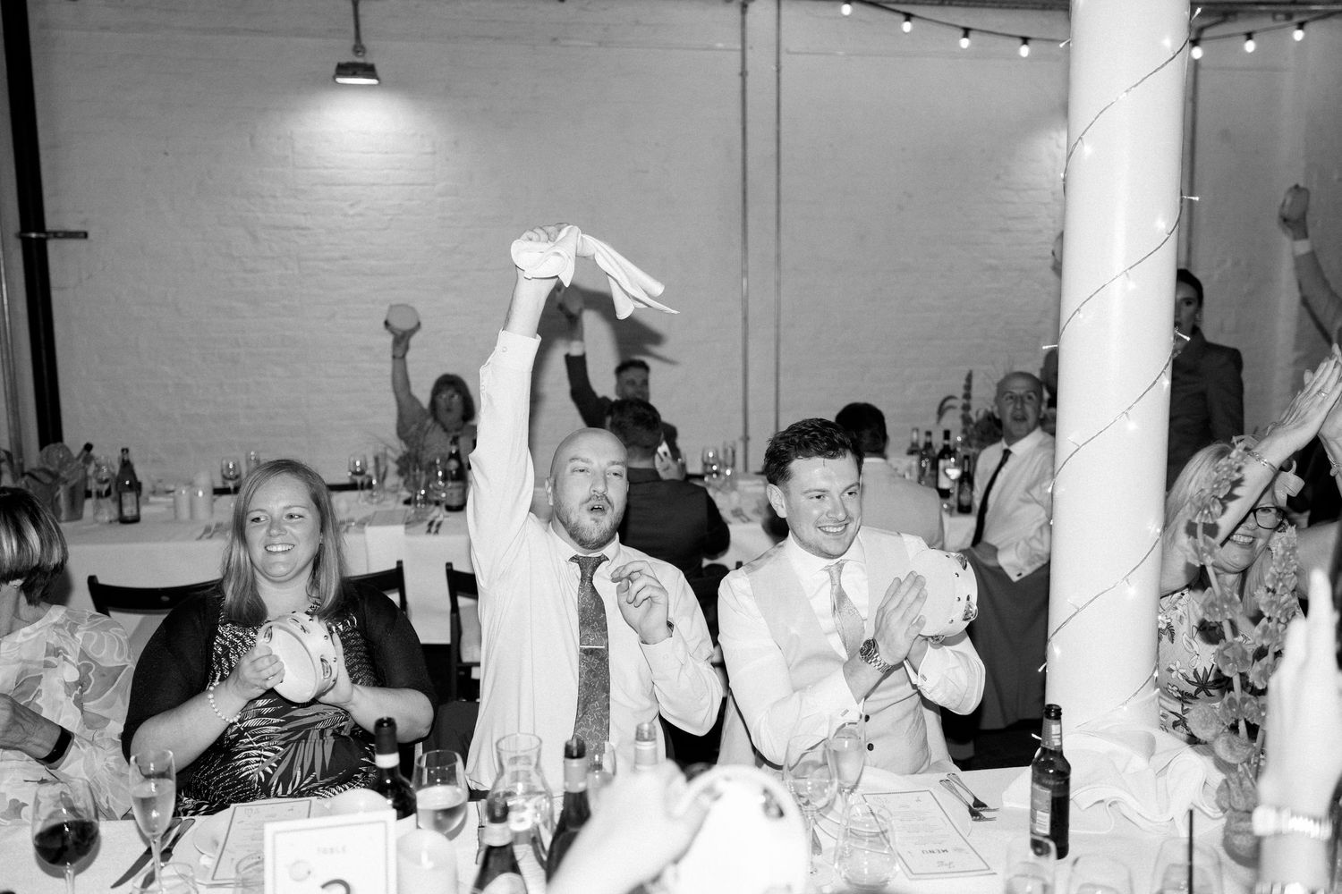 Wedding reception guests raise their hands in celebration under string lights.