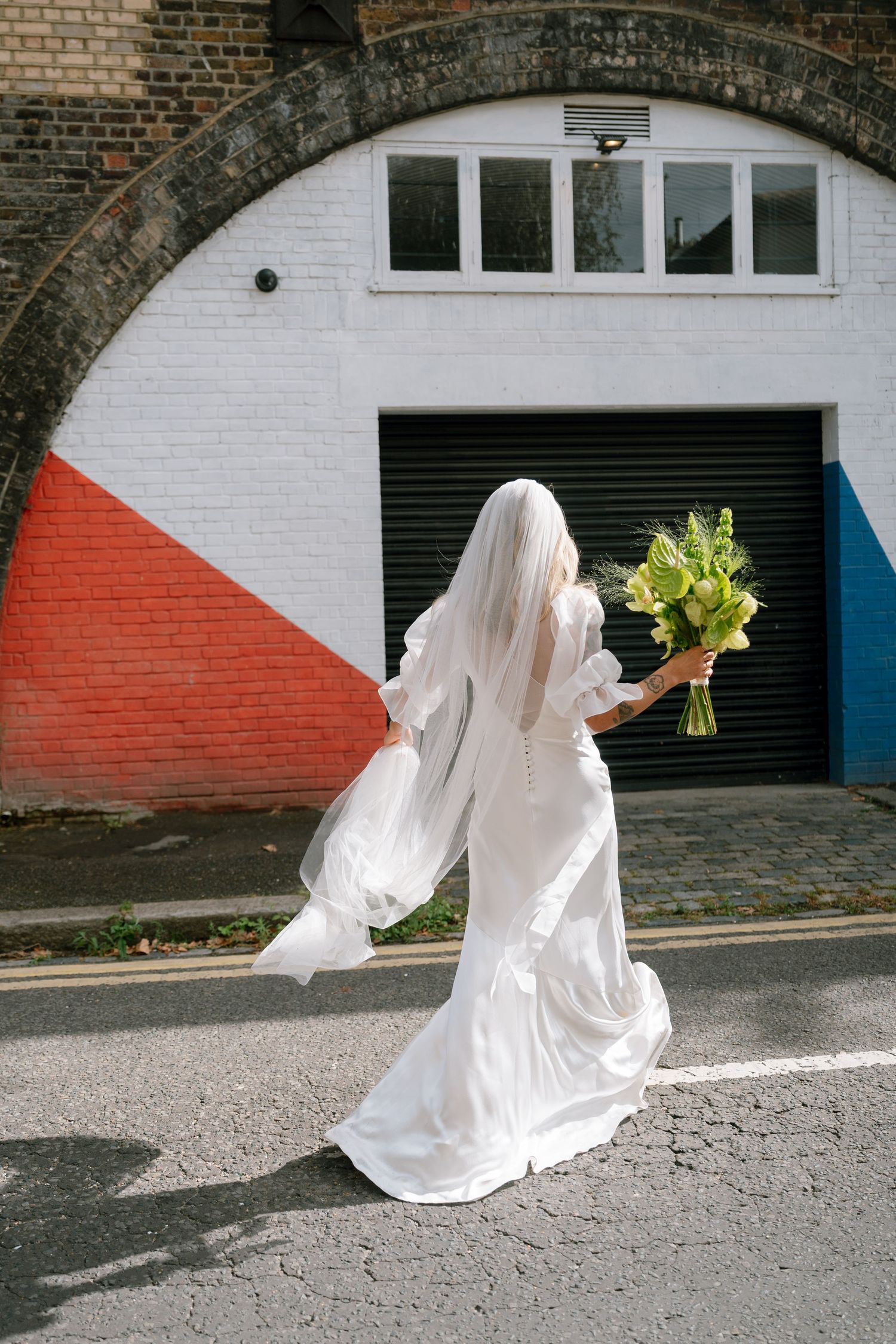 Person in flowing white dress poses dramatically against painted brick wall.