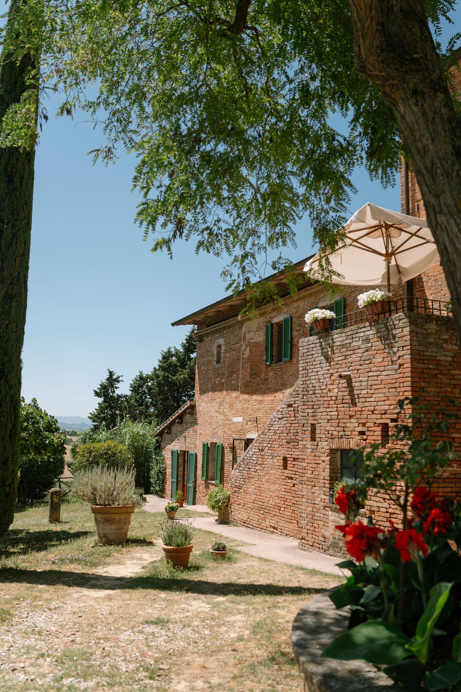 Rustic stone farmhouse buildings with red flowers in Tuscany surrounded by trees and gravel paths on a sunny day.