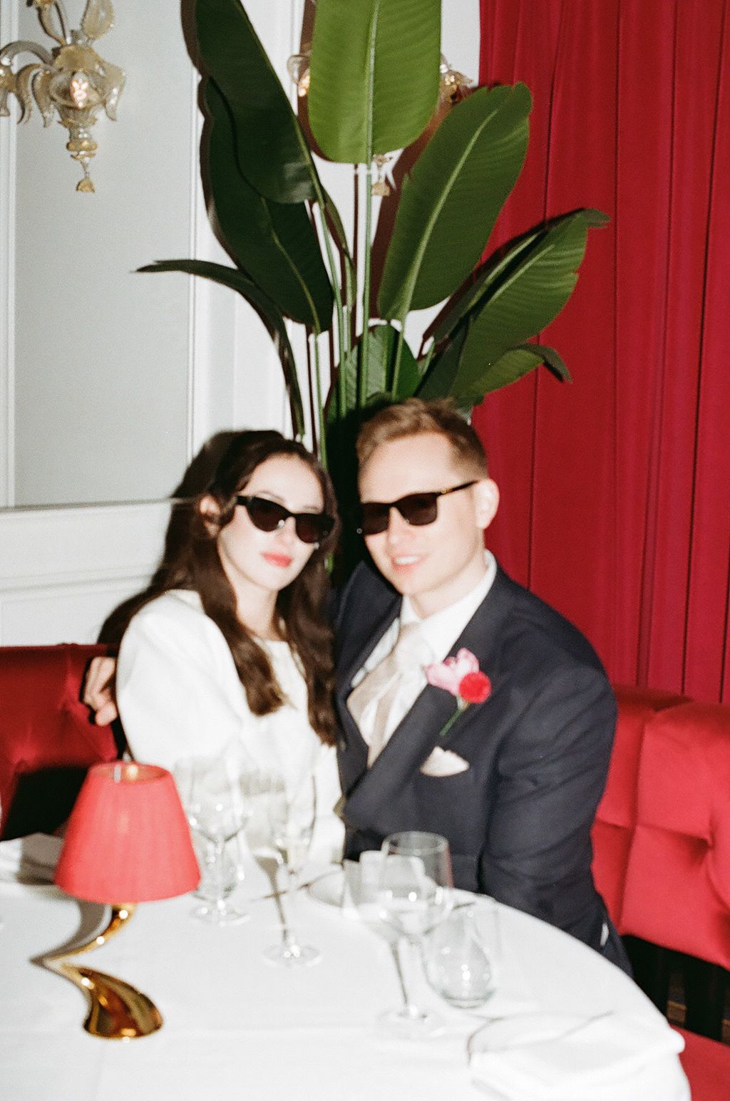 Two people in sunglasses sit at an elegant dining table with red decor and white tablecloths.