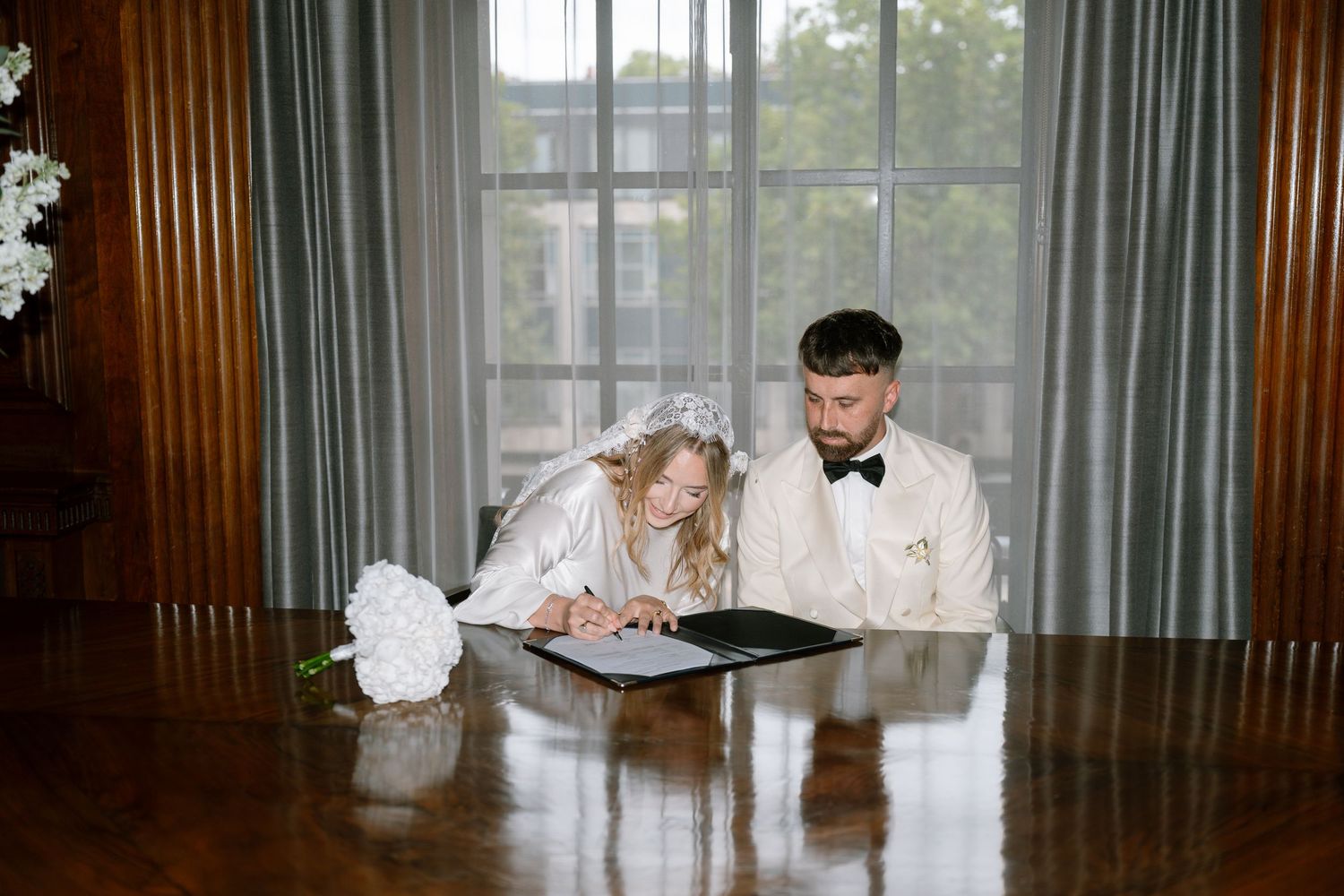 Wedding ceremony scene through a window with curtains and floral arrangements.