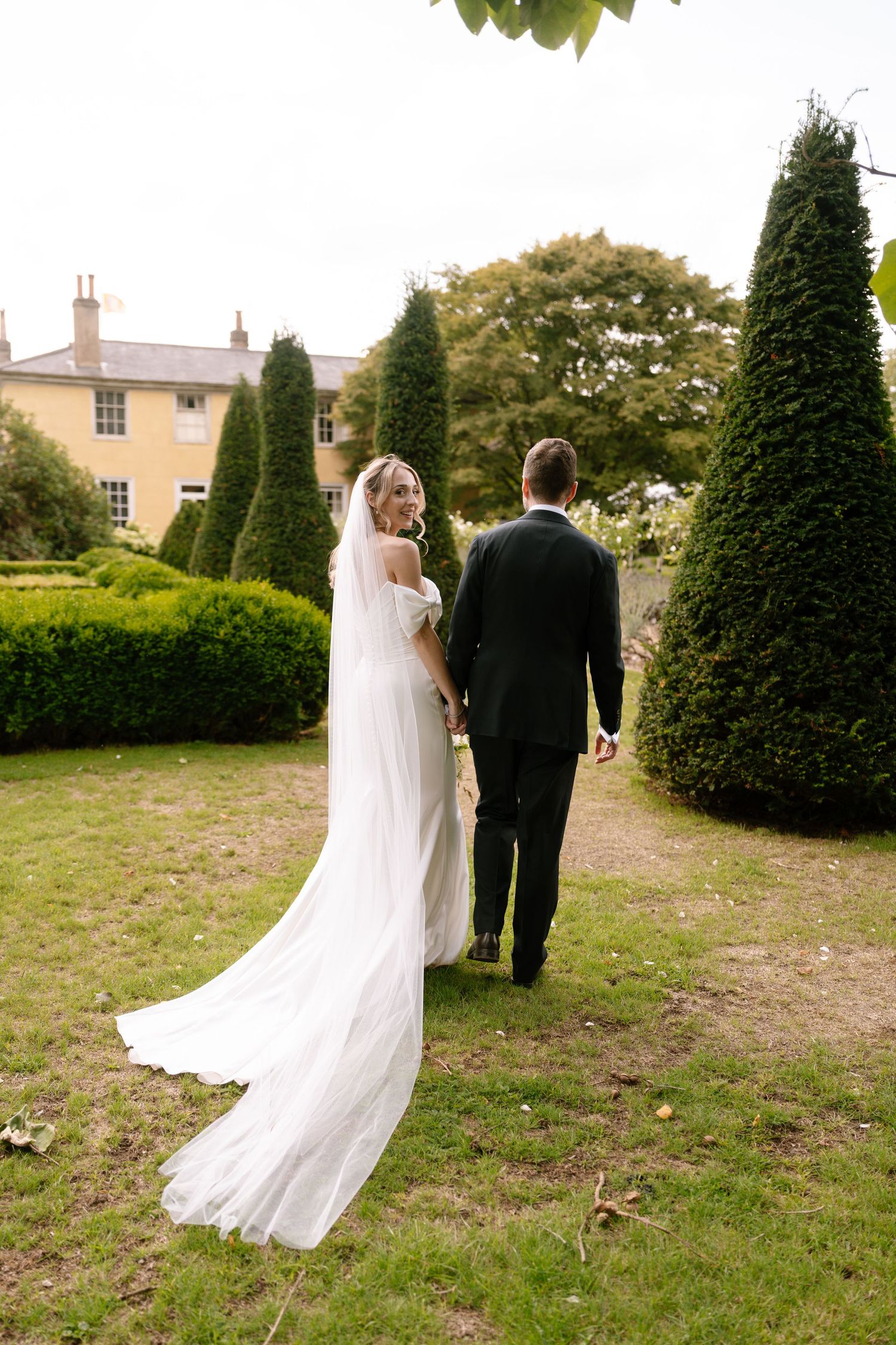 Newlyweds walk through formal garden with manicured topiary and historic building backdrop.