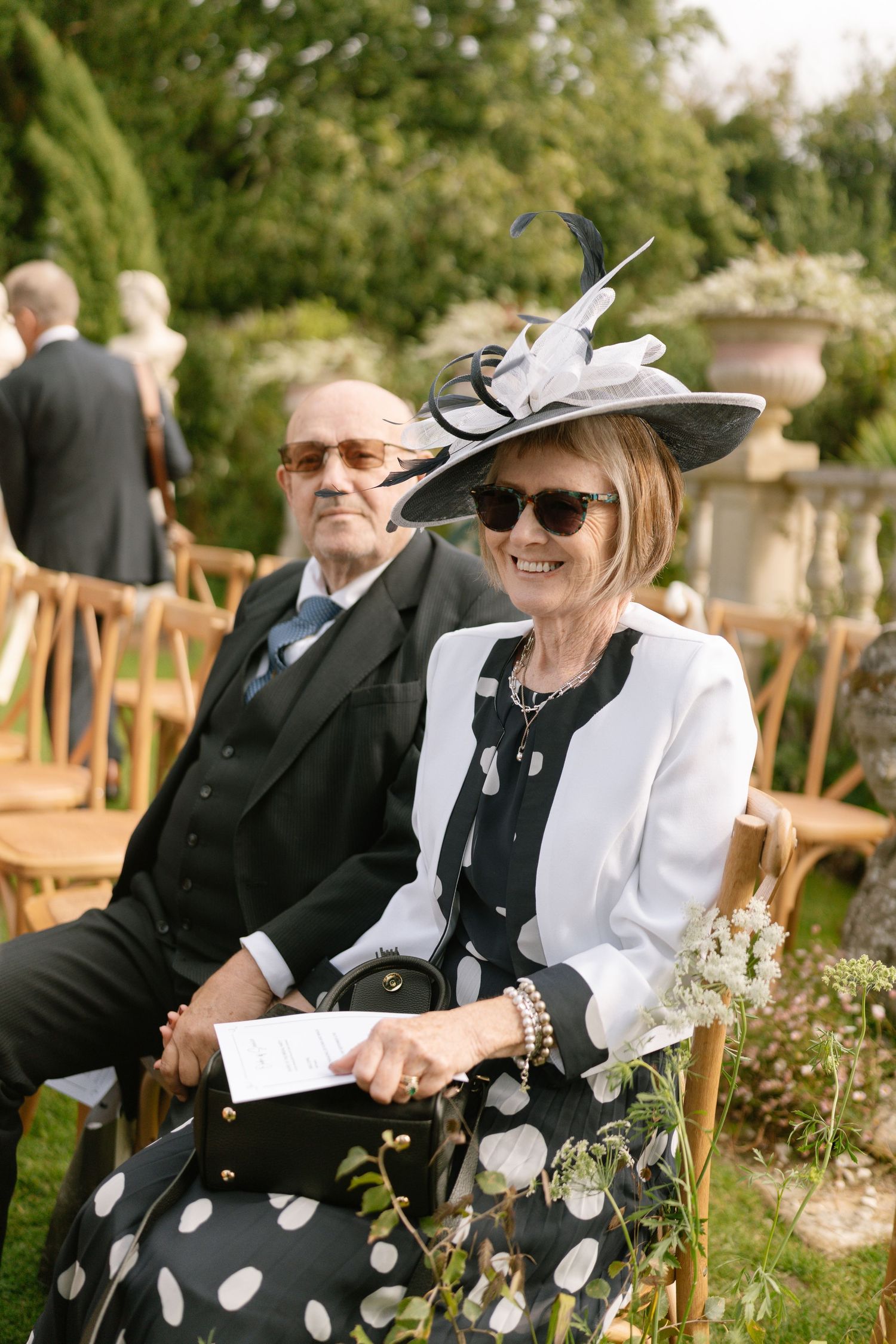 Elegantly dressed guests in formal attire and hats enjoying an outdoor wedding ceremony.