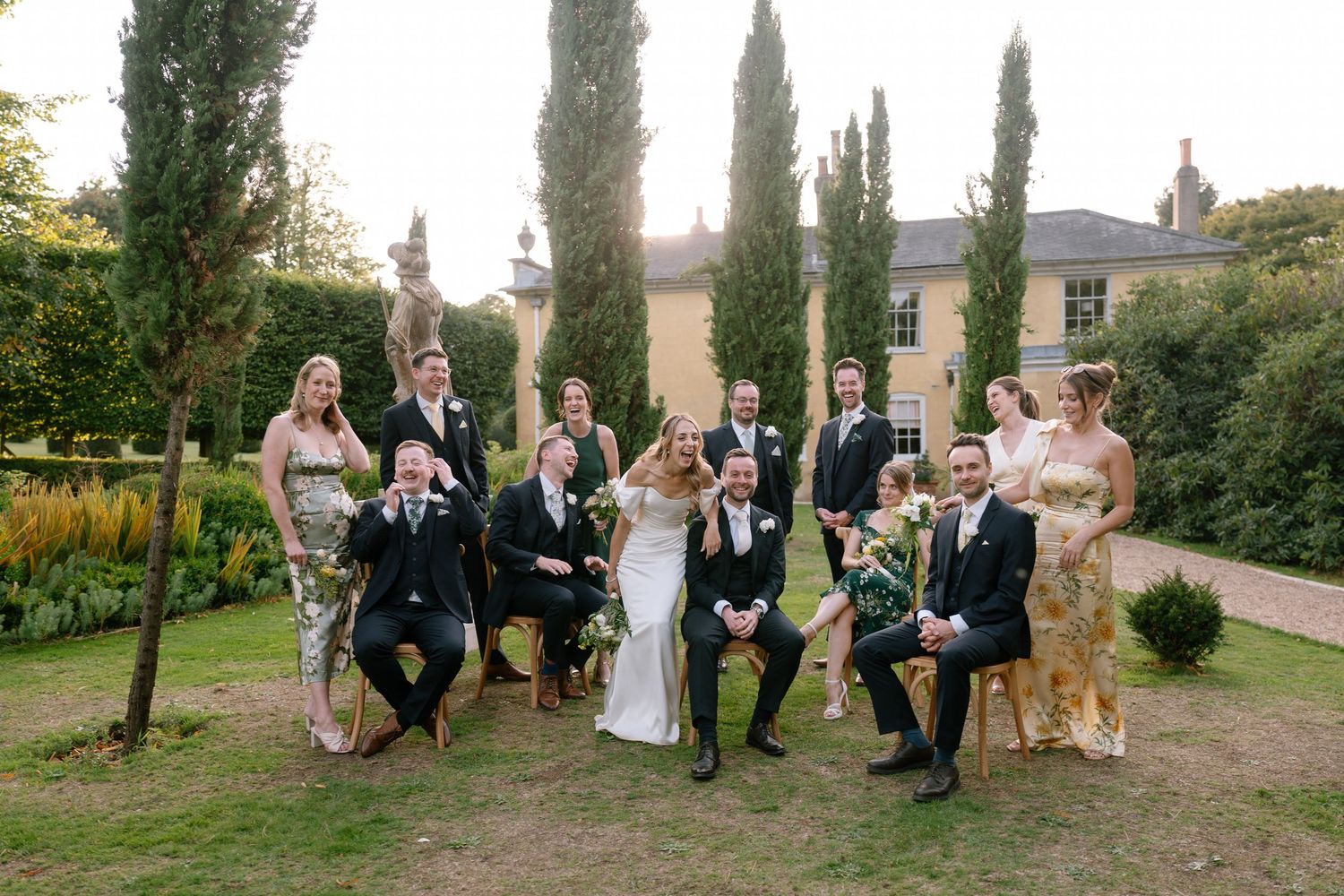 Wedding party and guests gathered for a formal group photo in a garden with tall cypress trees.