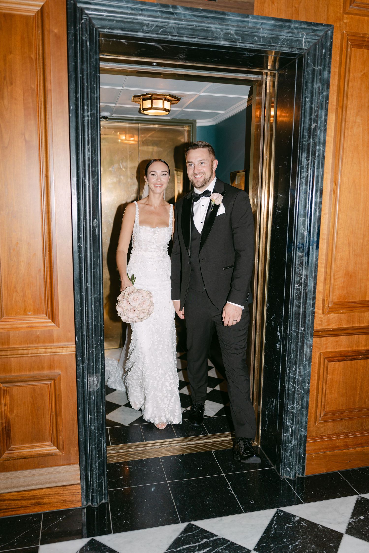 Newlyweds pose in a marble-framed elevator doorway at their wedding.