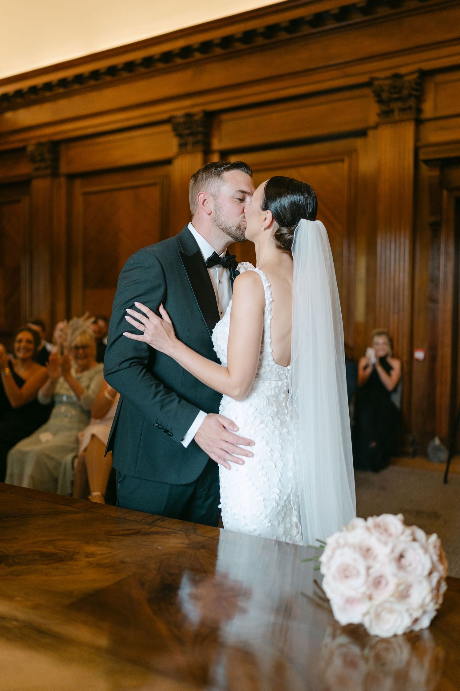 Newlyweds share a romantic moment in a warm-lit ballroom with wood paneling.