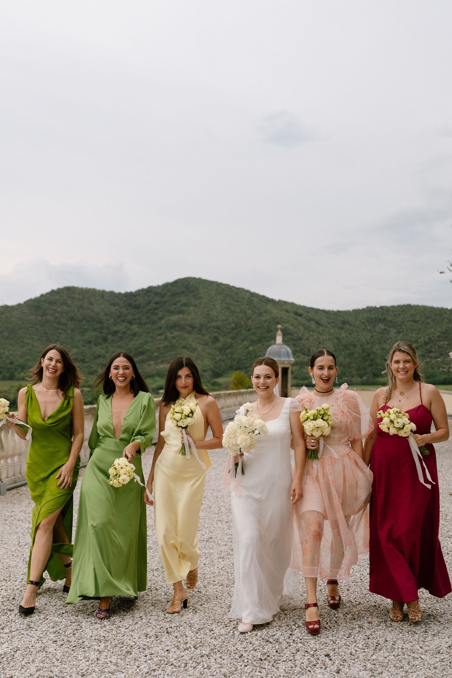 Wedding party in colorful dresses walks together on a gravel terrace with mountains in background.