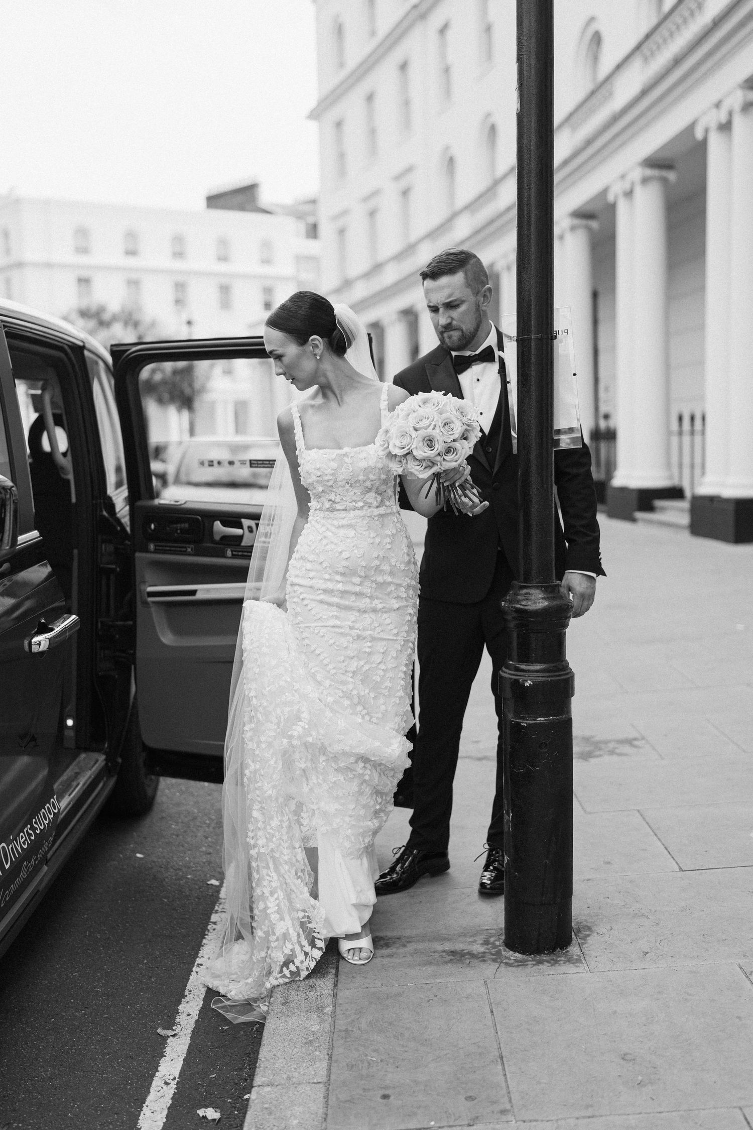 A couple exits a black car on their wedding day, with the bride in a lace gown holding a white bouquet.