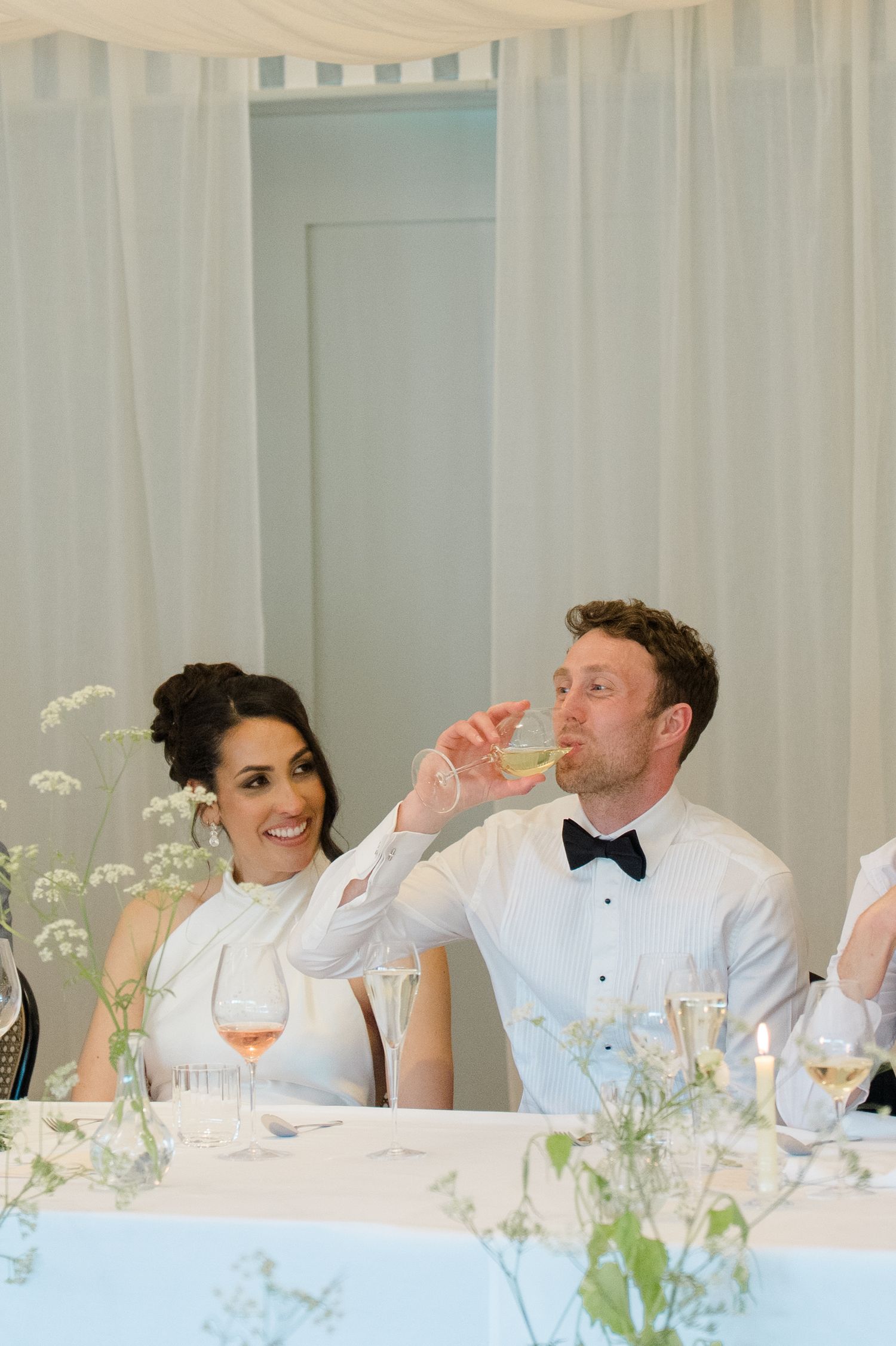 Bride smiles as groom takes a shot at elegant wedding reception table with floral centerpieces.