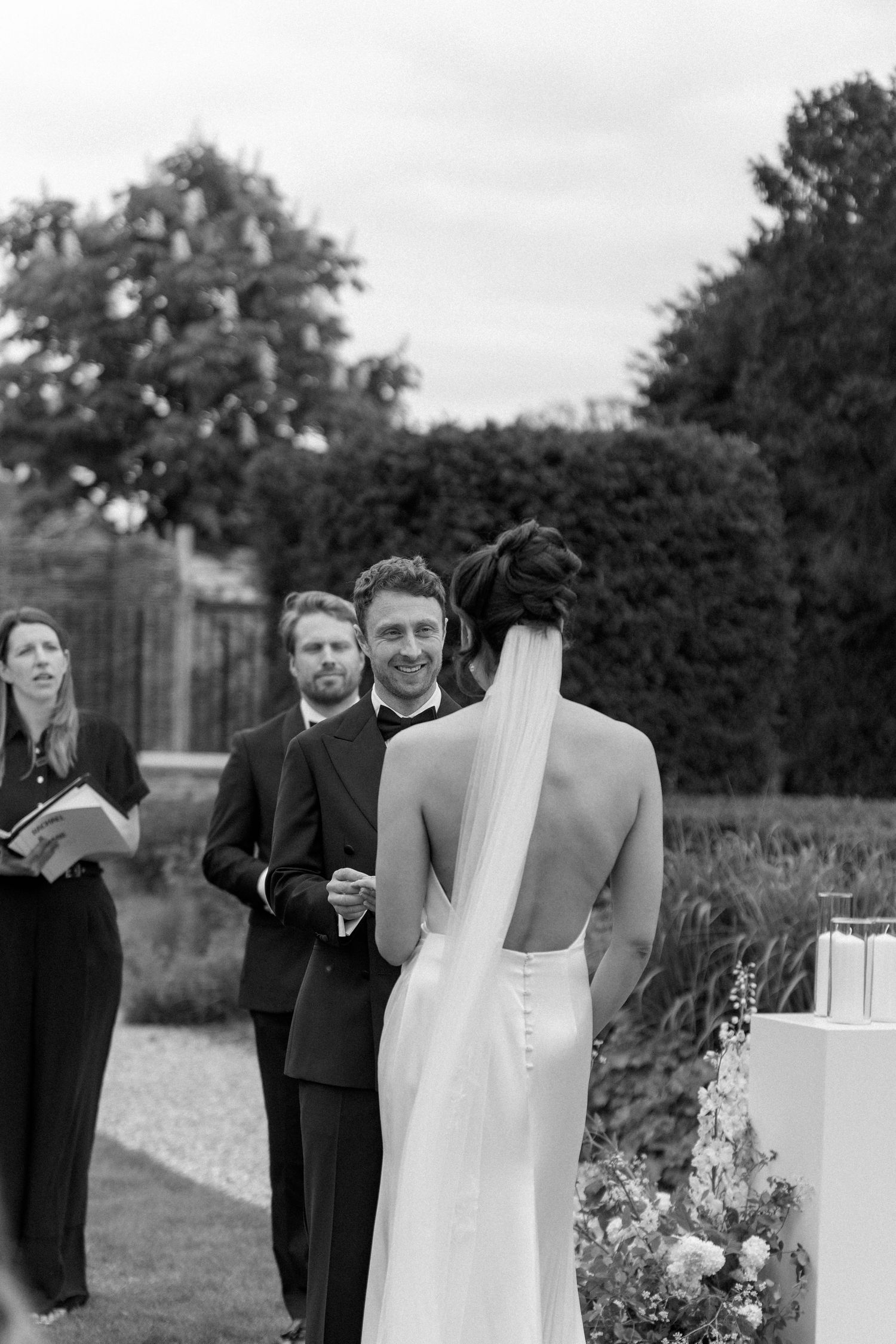 Groom smiling at bride during outdoor wedding ceremony with groomsman and officiant in background.