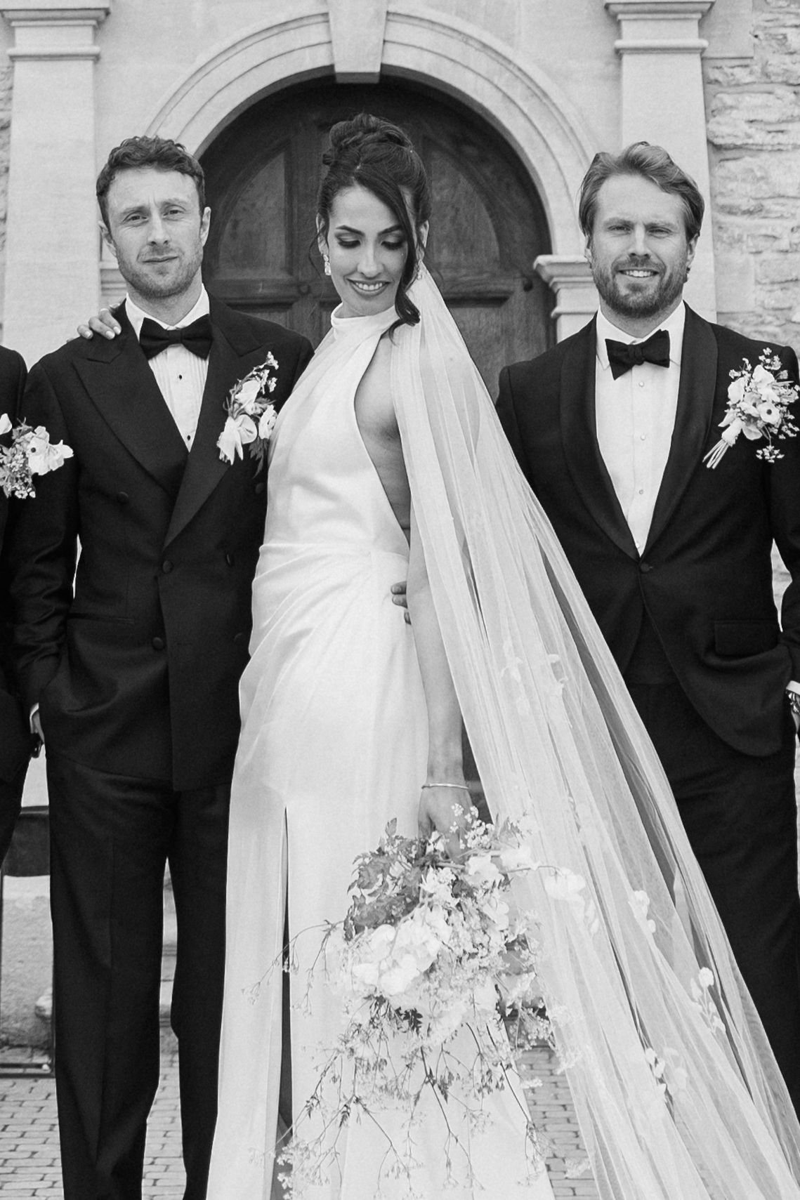 Black and white portrait of bride in halter gown with veil flanked by two groomsmen in tuxedos.