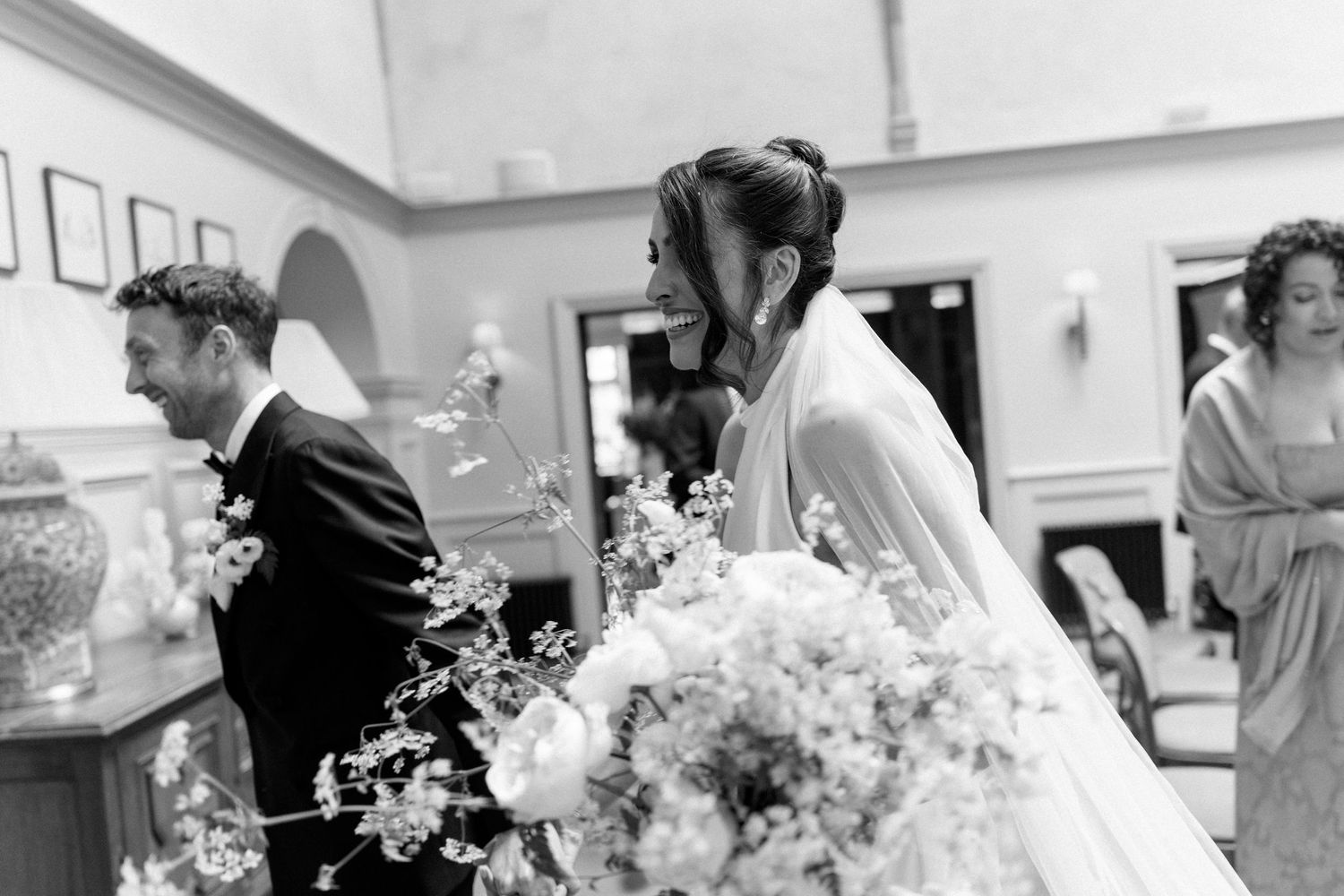 Black and white photo of bride holding floral bouquet with groom during elegant indoor wedding ceremony.