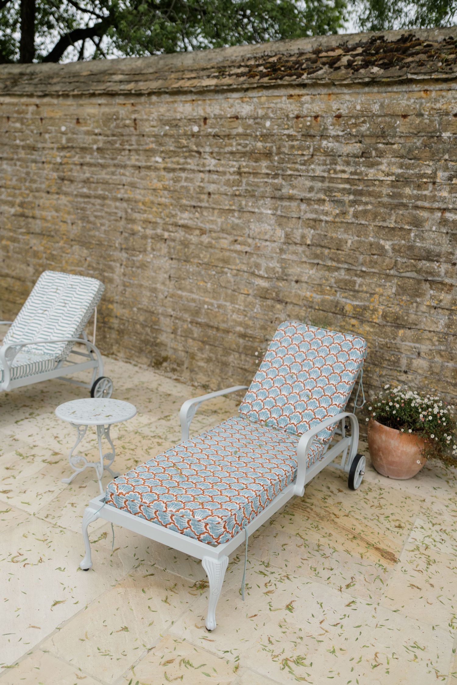 Two white lounge chairs with colorful cushions on stone patio against rustic brick wall.
