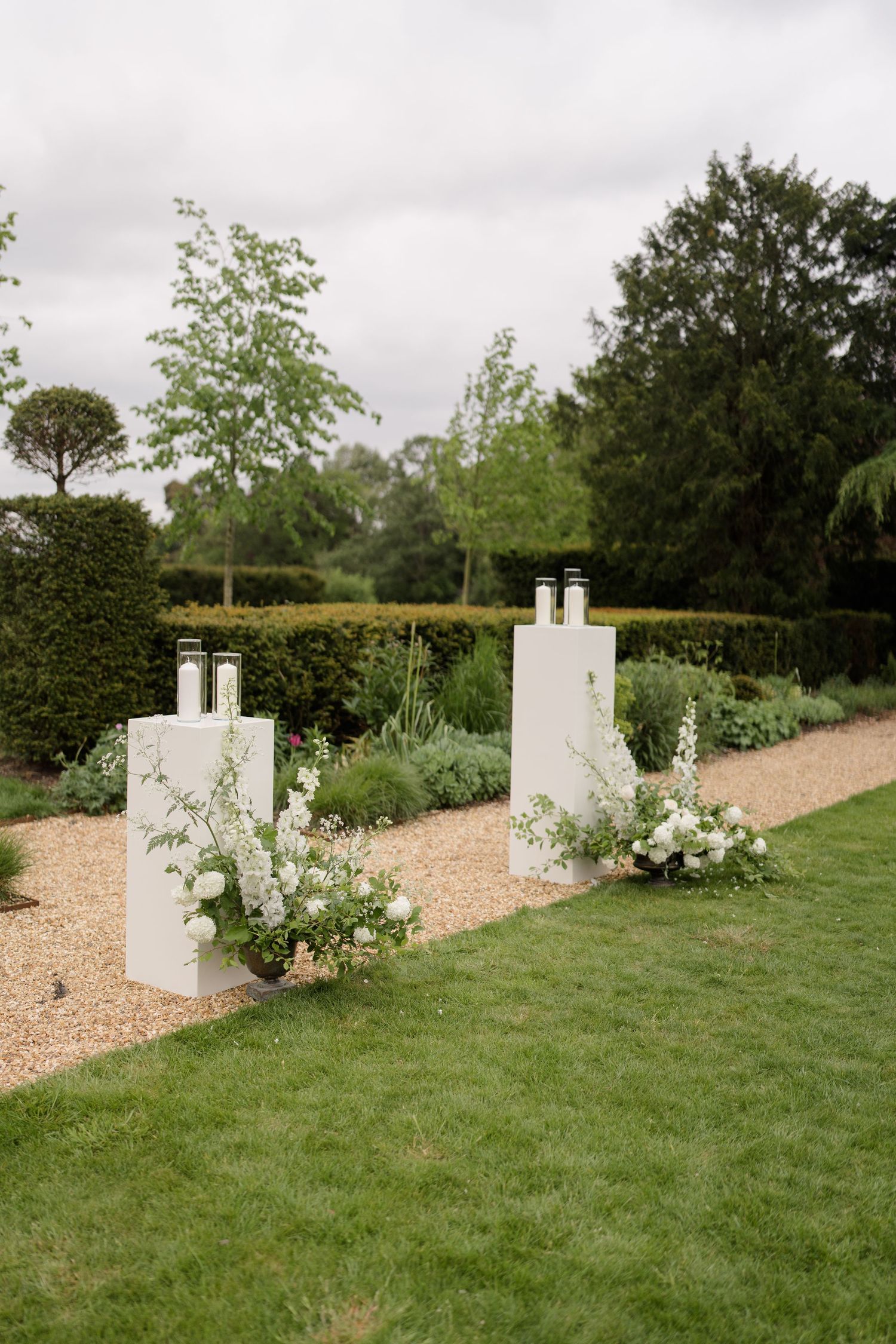 White wedding ceremony pedestals with black lanterns and white floral arrangements on a gravel path in a formal garden.