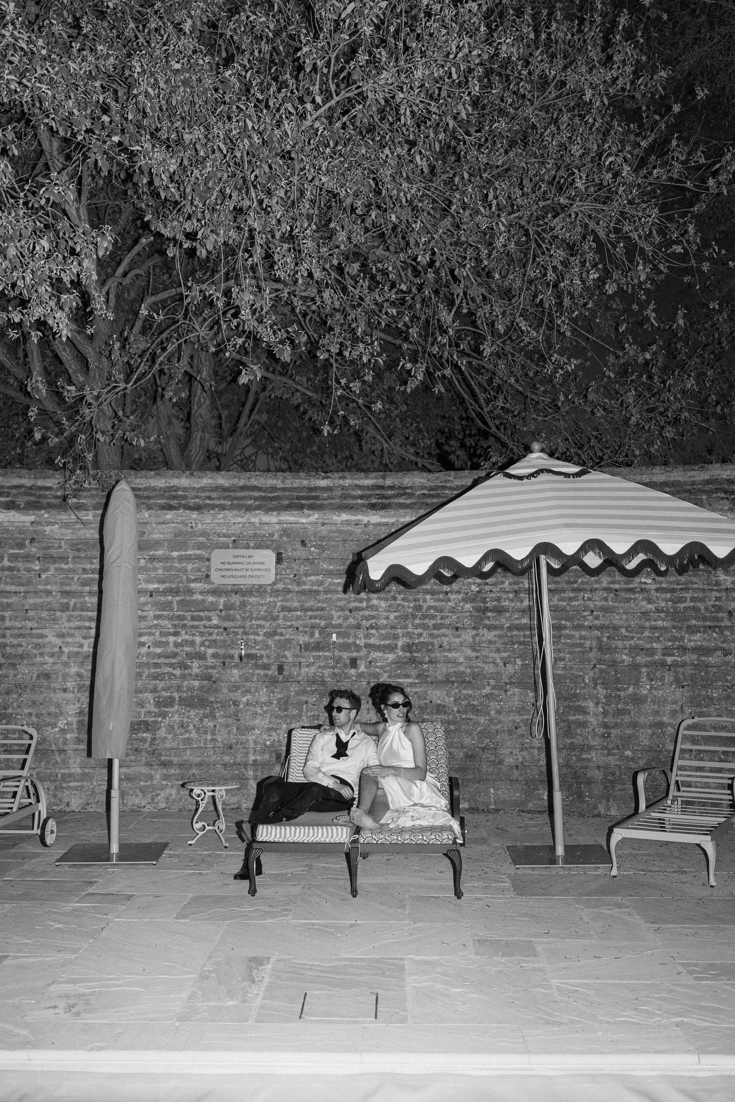 Newlyweds in sunglasses relax on bench under striped umbrella by brick wall at night in black and white.