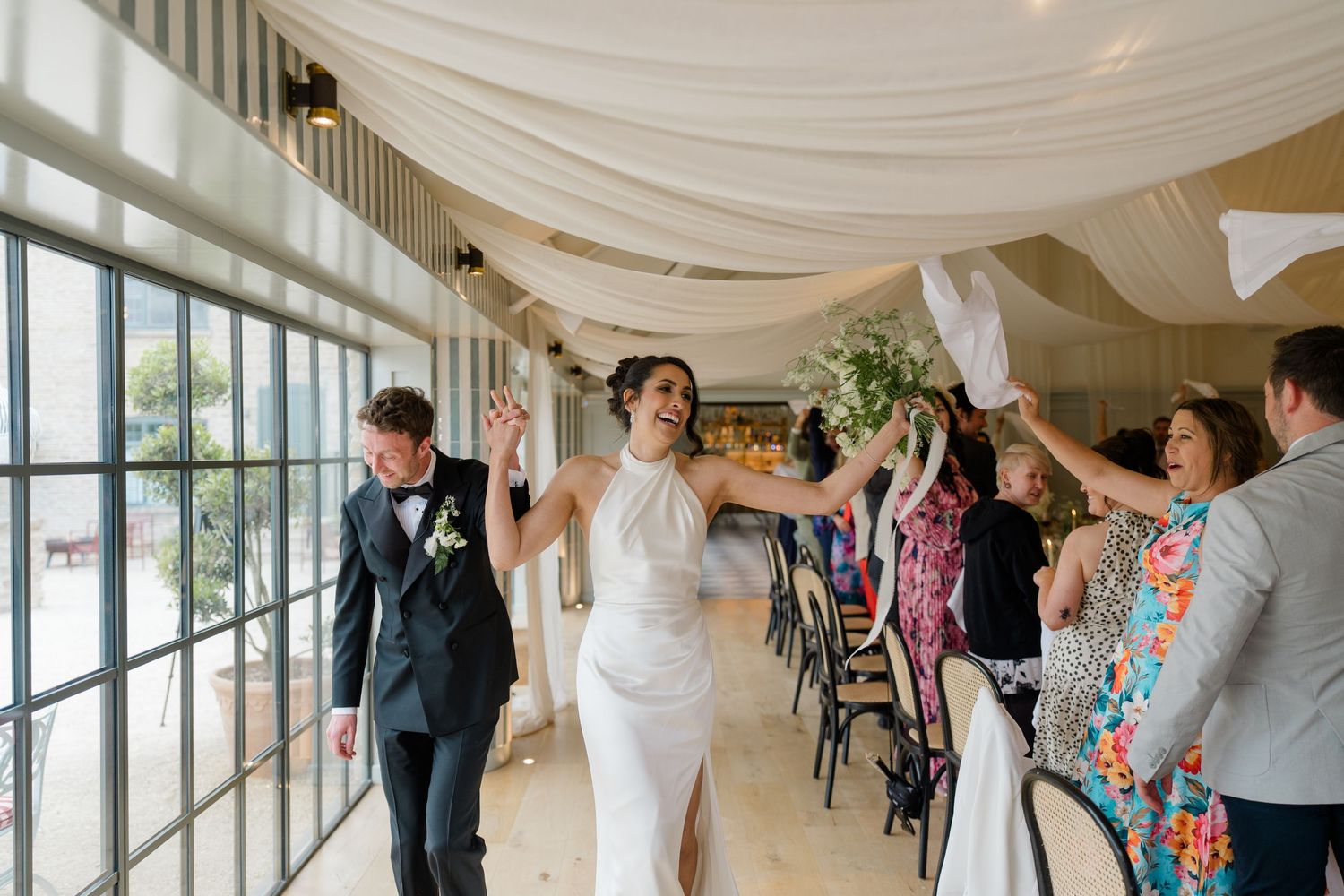 Bride and groom make joyful entrance into wedding reception as guests wave napkins in celebration.