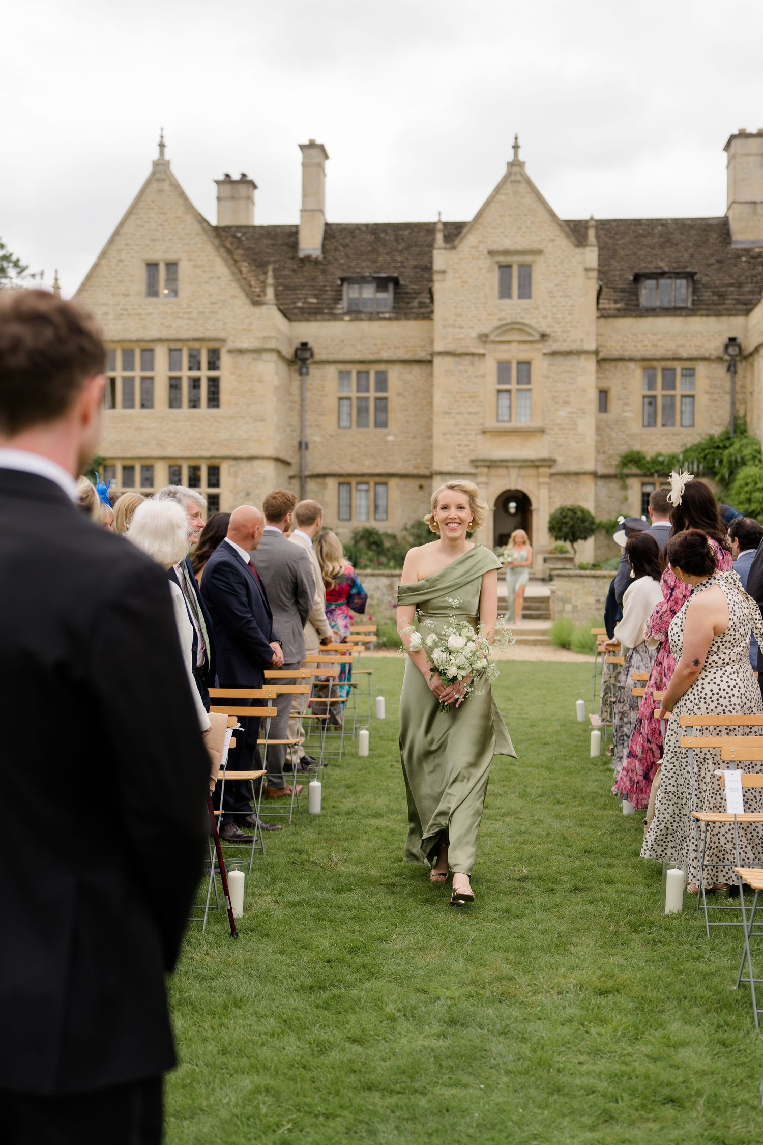 Bridesmaid in sage green gown walks down outdoor aisle at English manor house wedding ceremony.