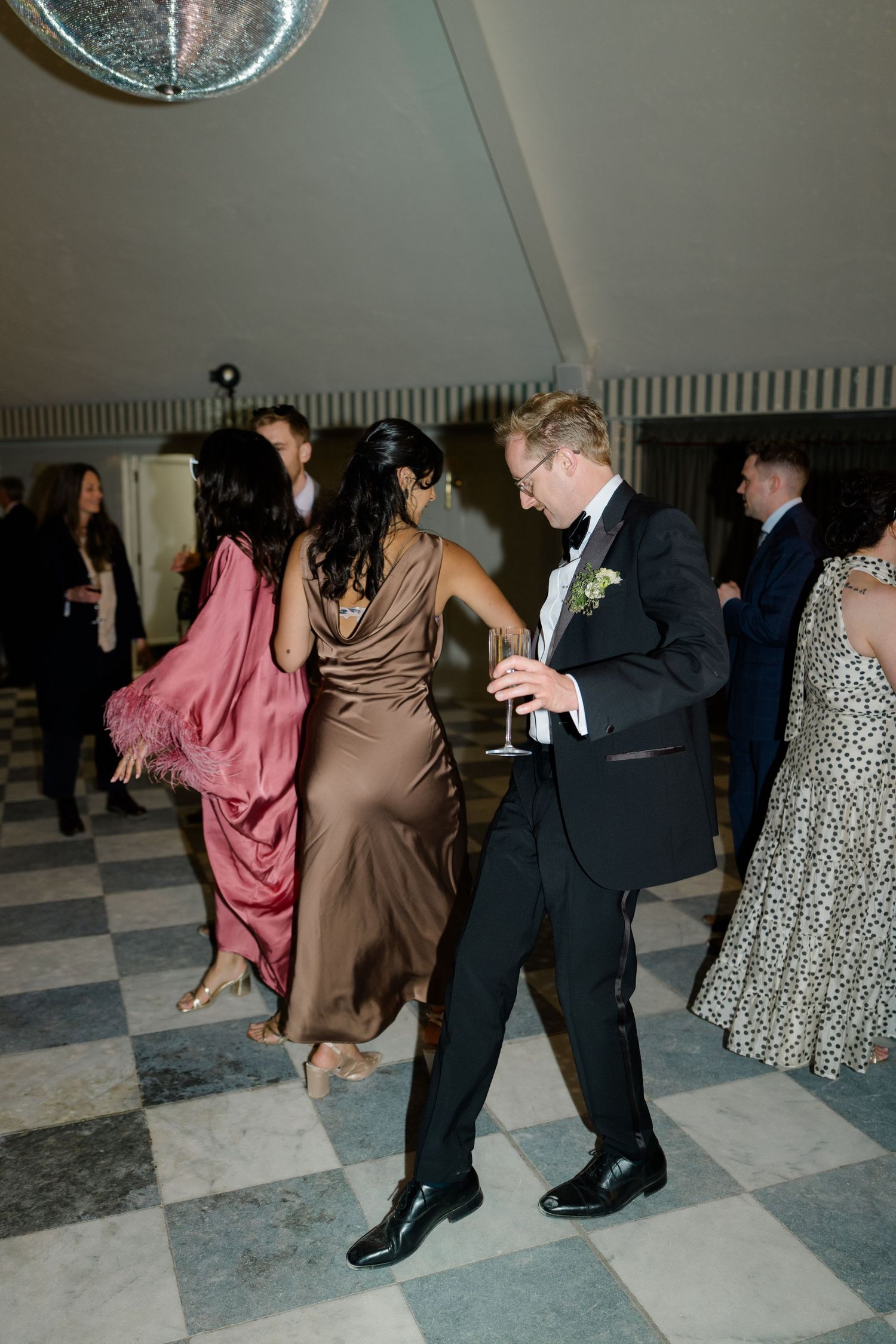 Guests dancing on checkered floor at elegant wedding reception under disco ball with colorful gowns.