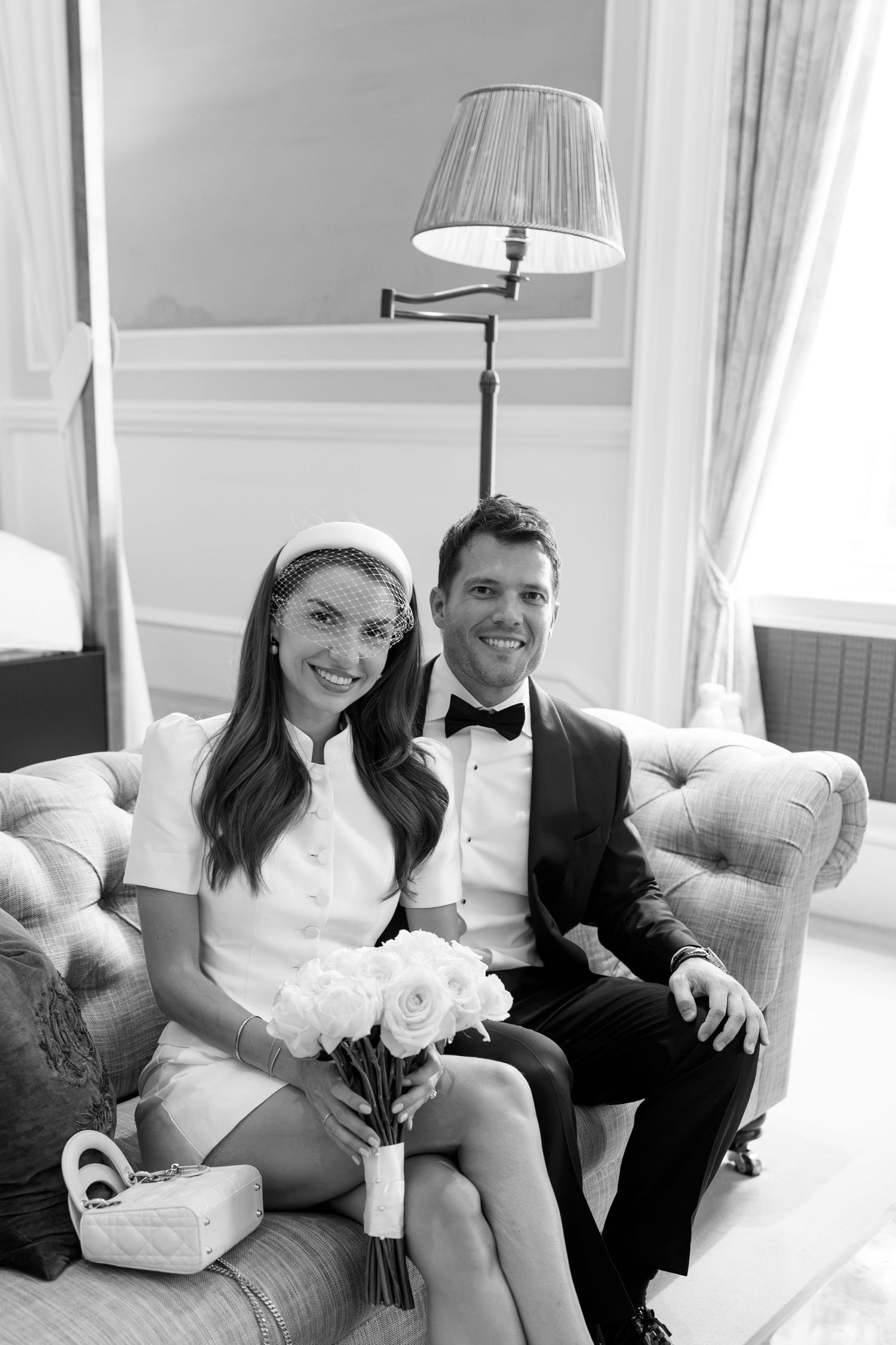 Bride and groom sitting on elegant sofa, she holds white roses bouquet wearing mini dress and veil, he wears tuxedo.