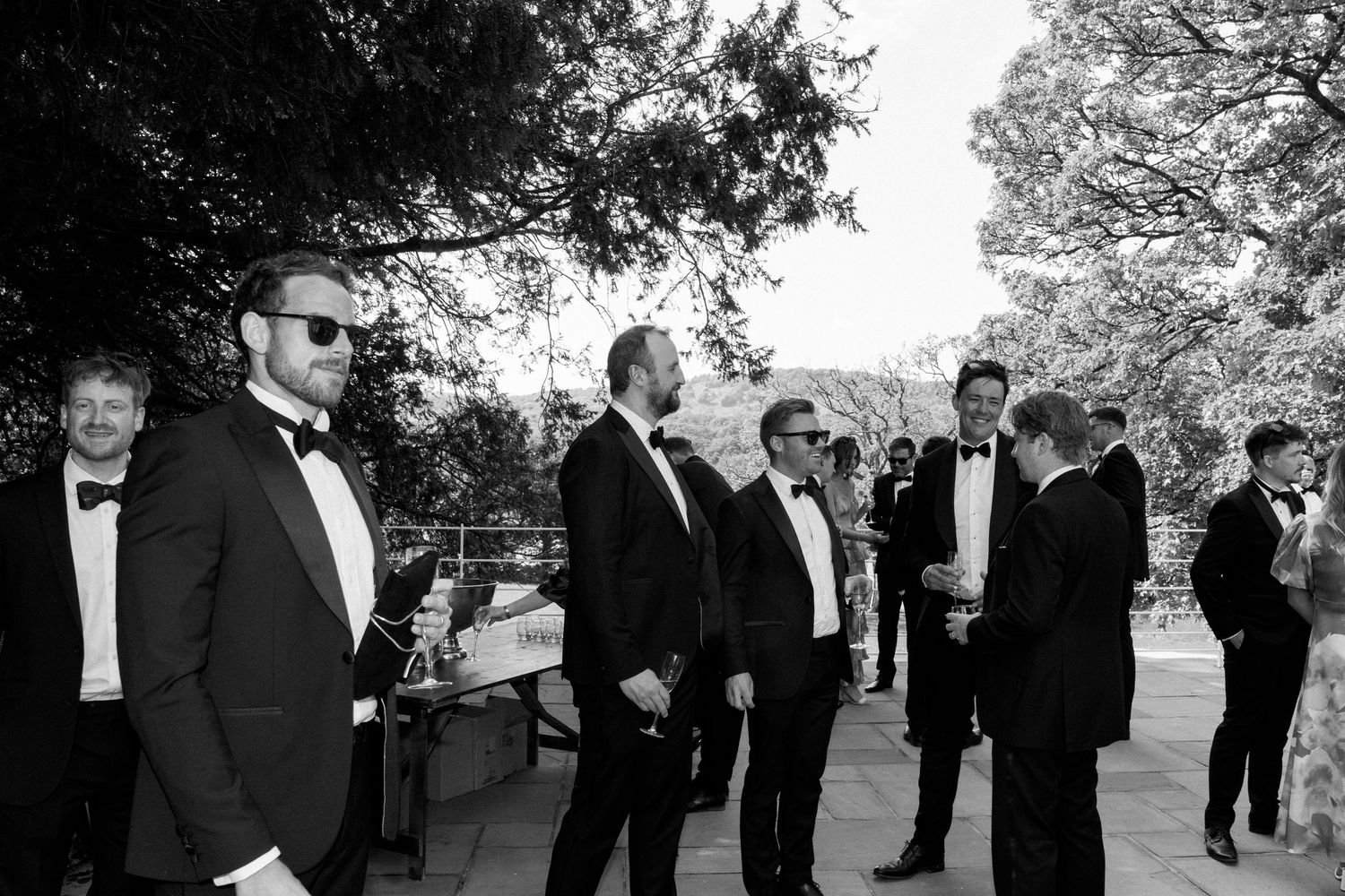 A group in formal black attire stands together outdoors during a wedding celebration.