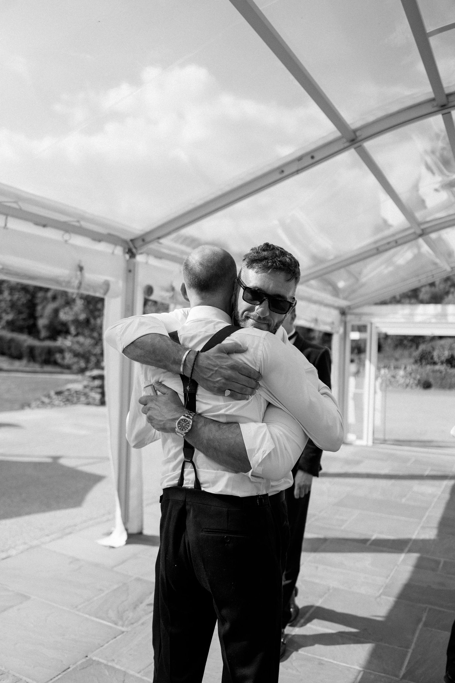 Black and white photo of wedding party members embracing outdoors under a tent structure.