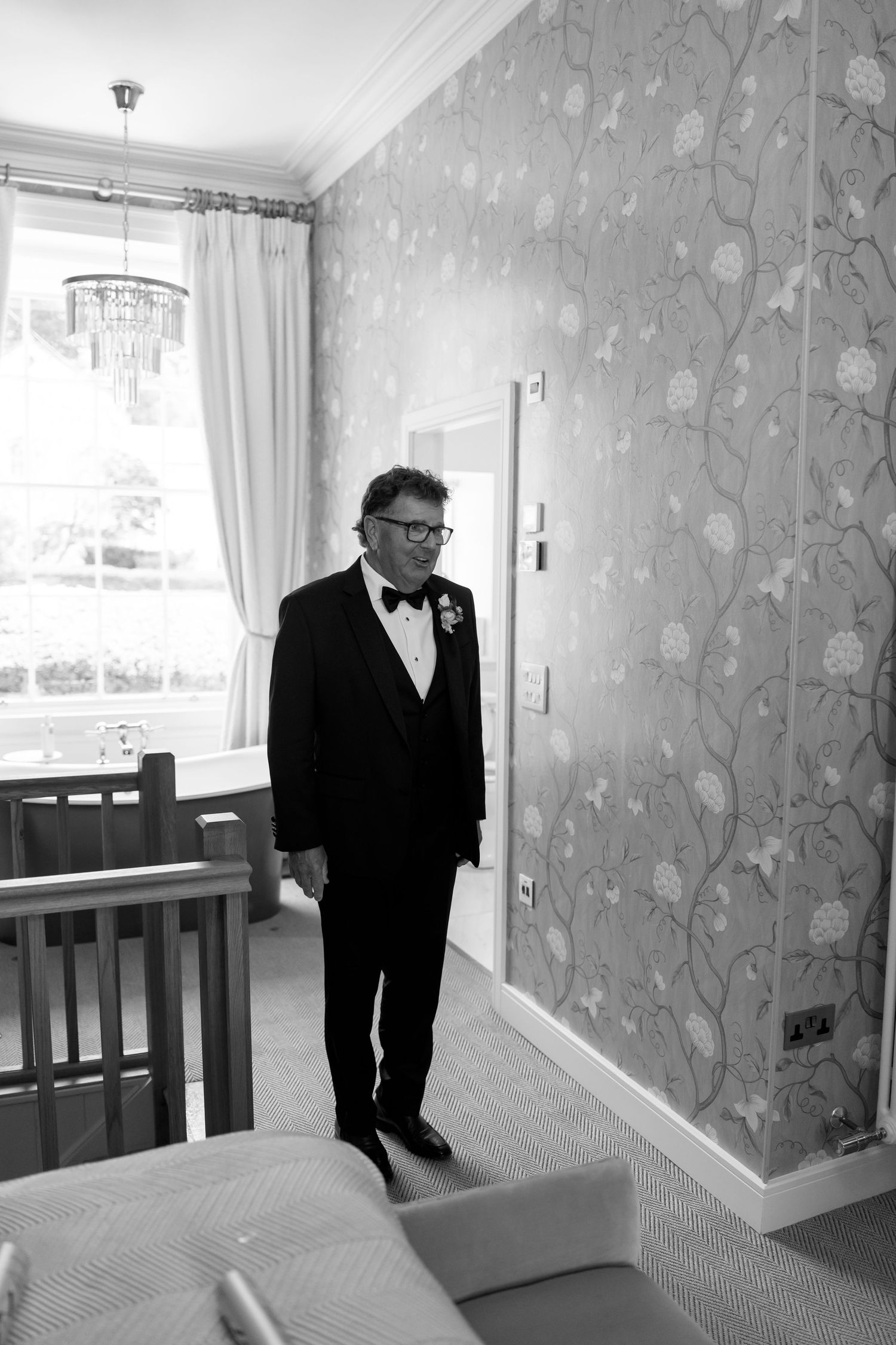 A formal black and white photo of someone in a tuxedo standing in an elegantly wallpapered room.