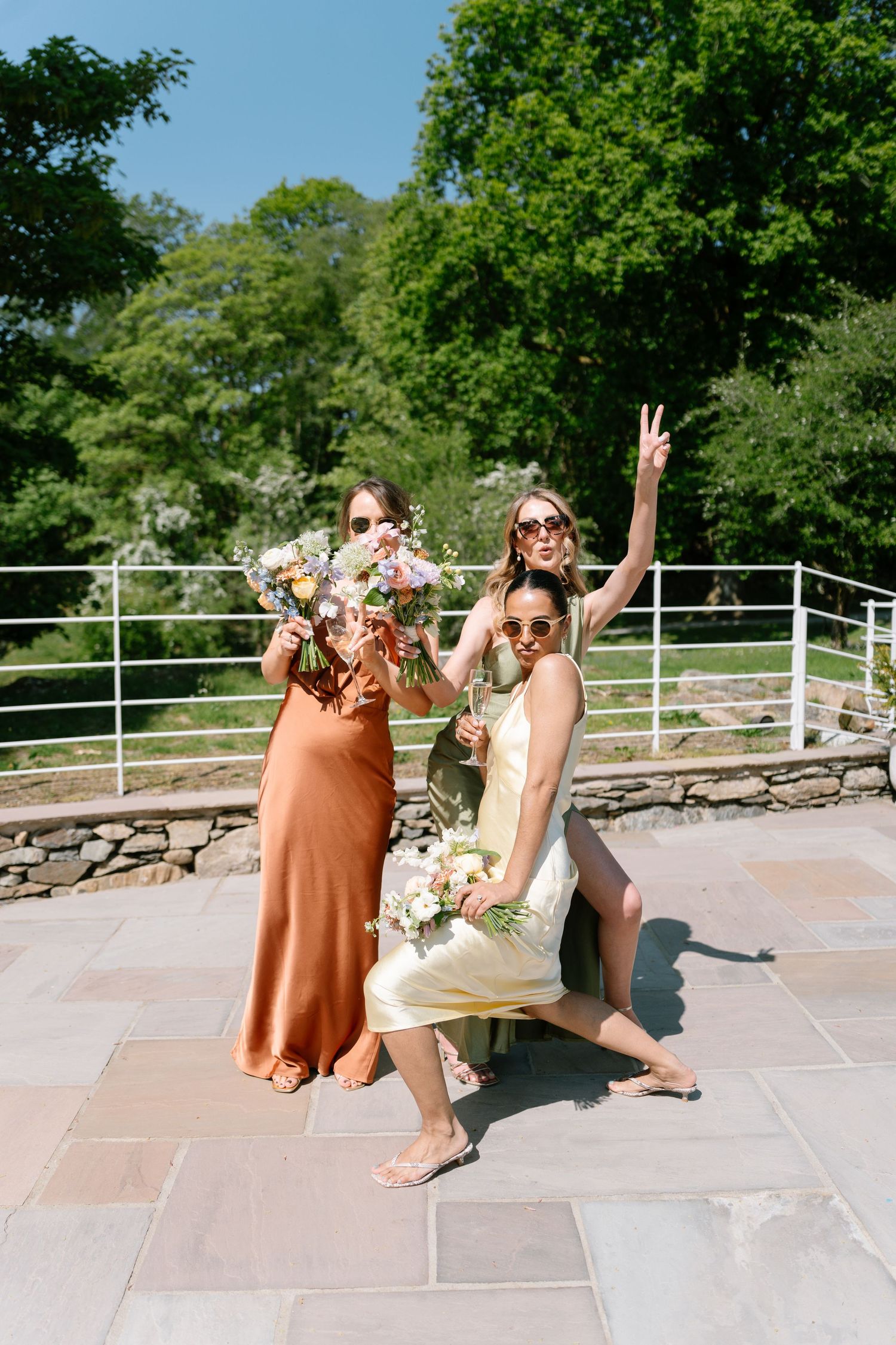 Bridesmaids in orange dresses pose playfully on a stone patio with white railing.