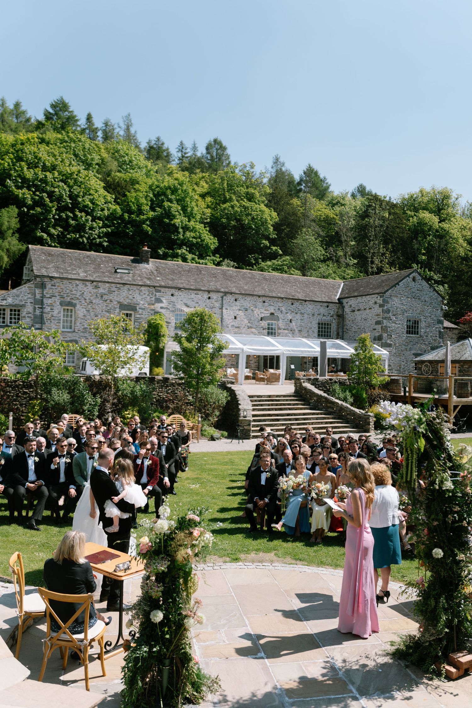 Outdoor wedding ceremony setup with stone buildings, floral decorations, and guests seated on wooden chairs.