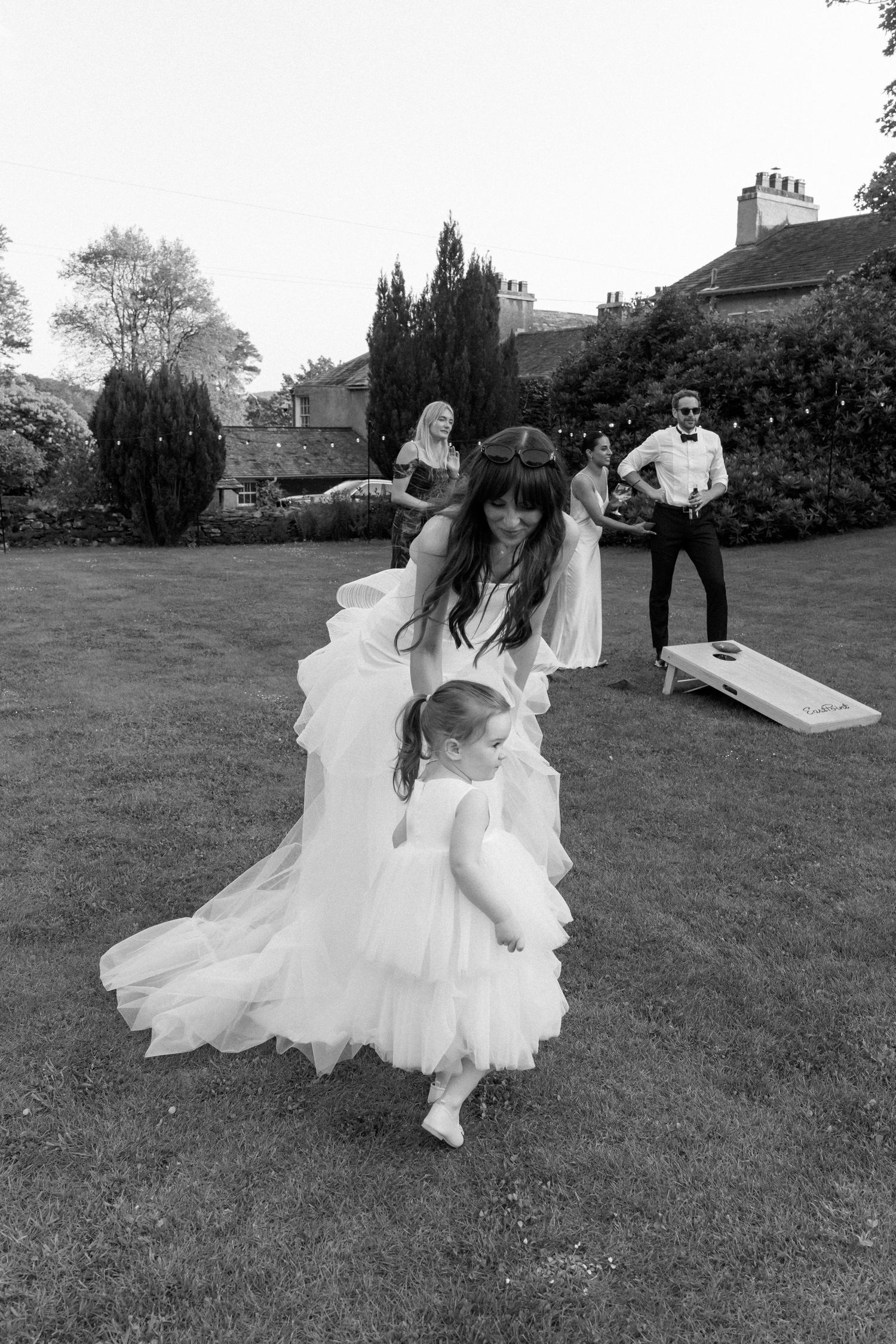 Wedding party members in formal attire play on a lawn with a white dress flowing in the breeze.