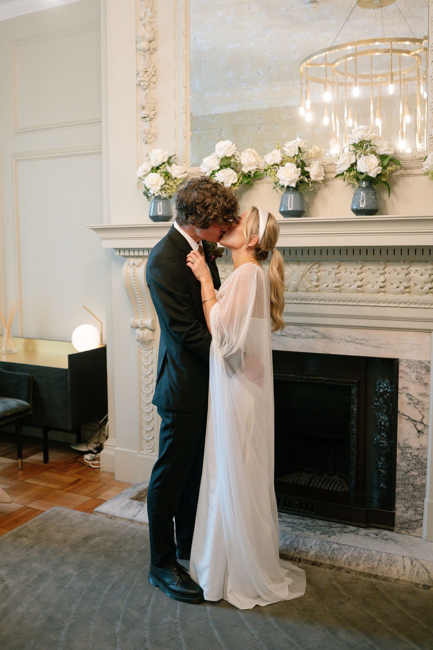 A romantic first kiss sequence between newlyweds in front of an elegant marble fireplace with floral decorations.