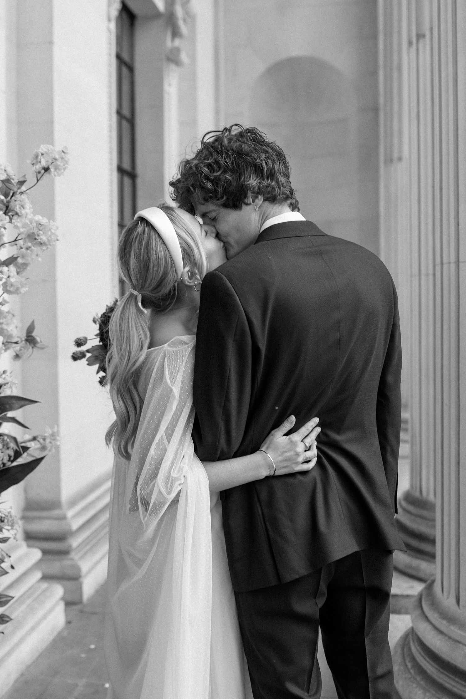 A black and white wedding photo showing a couple embracing between elegant columns.