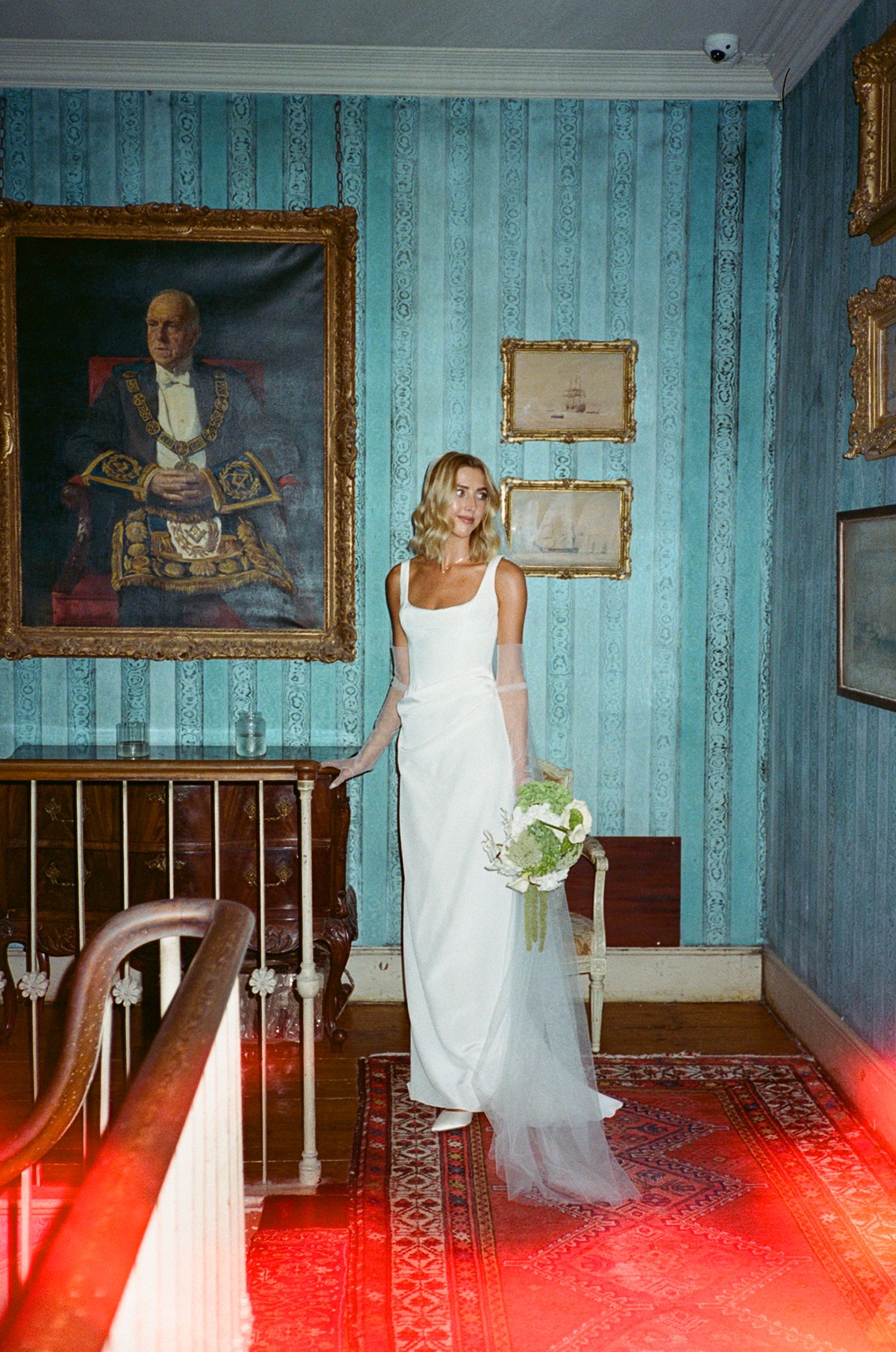 Bride in white gown holding bouquet stands before blue wallpaper and portrait painting in elegant historic venue.