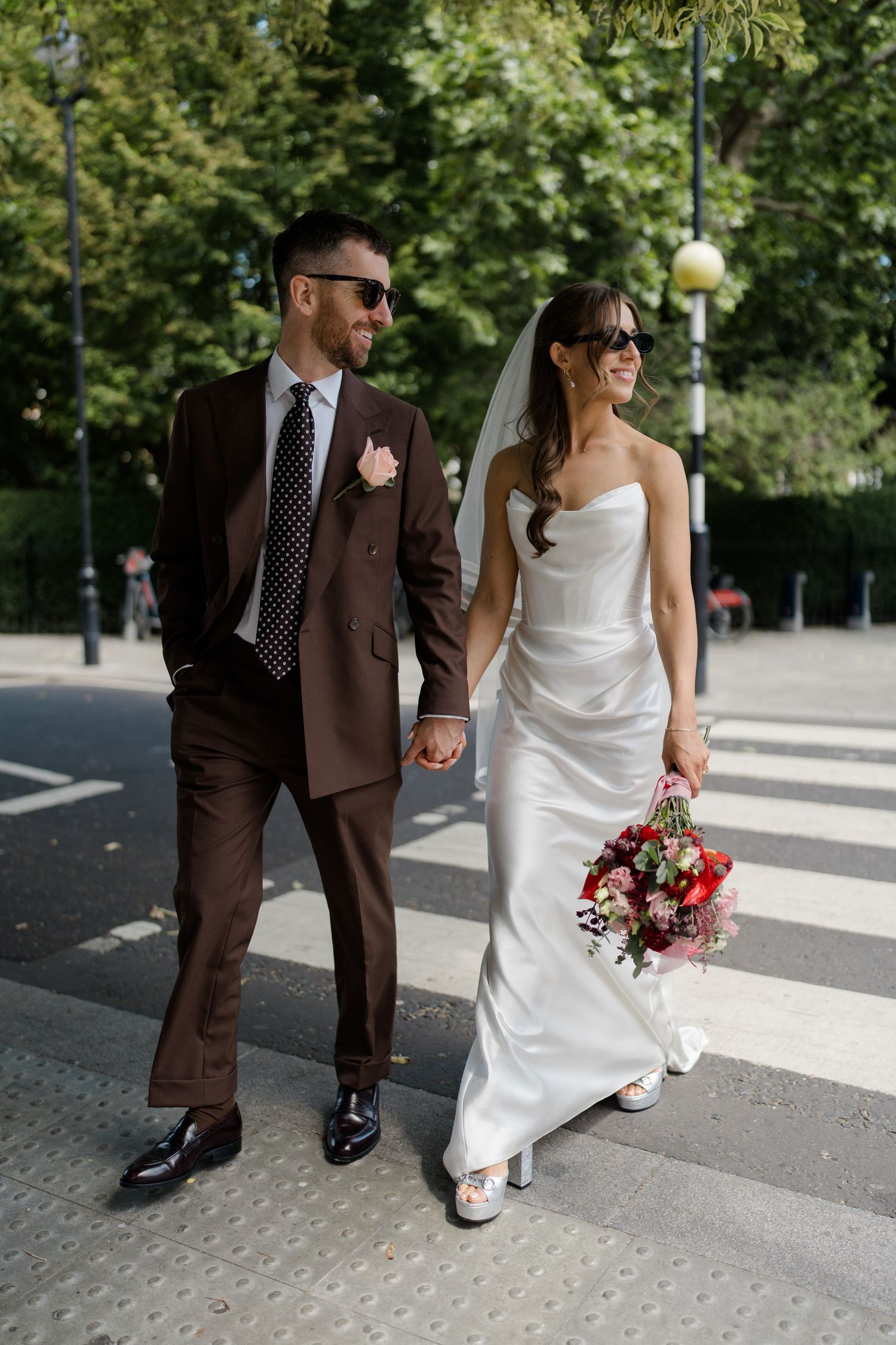 Bride and groom in sunglasses crossing a zebra crossing holding hands with red bridal bouquet.