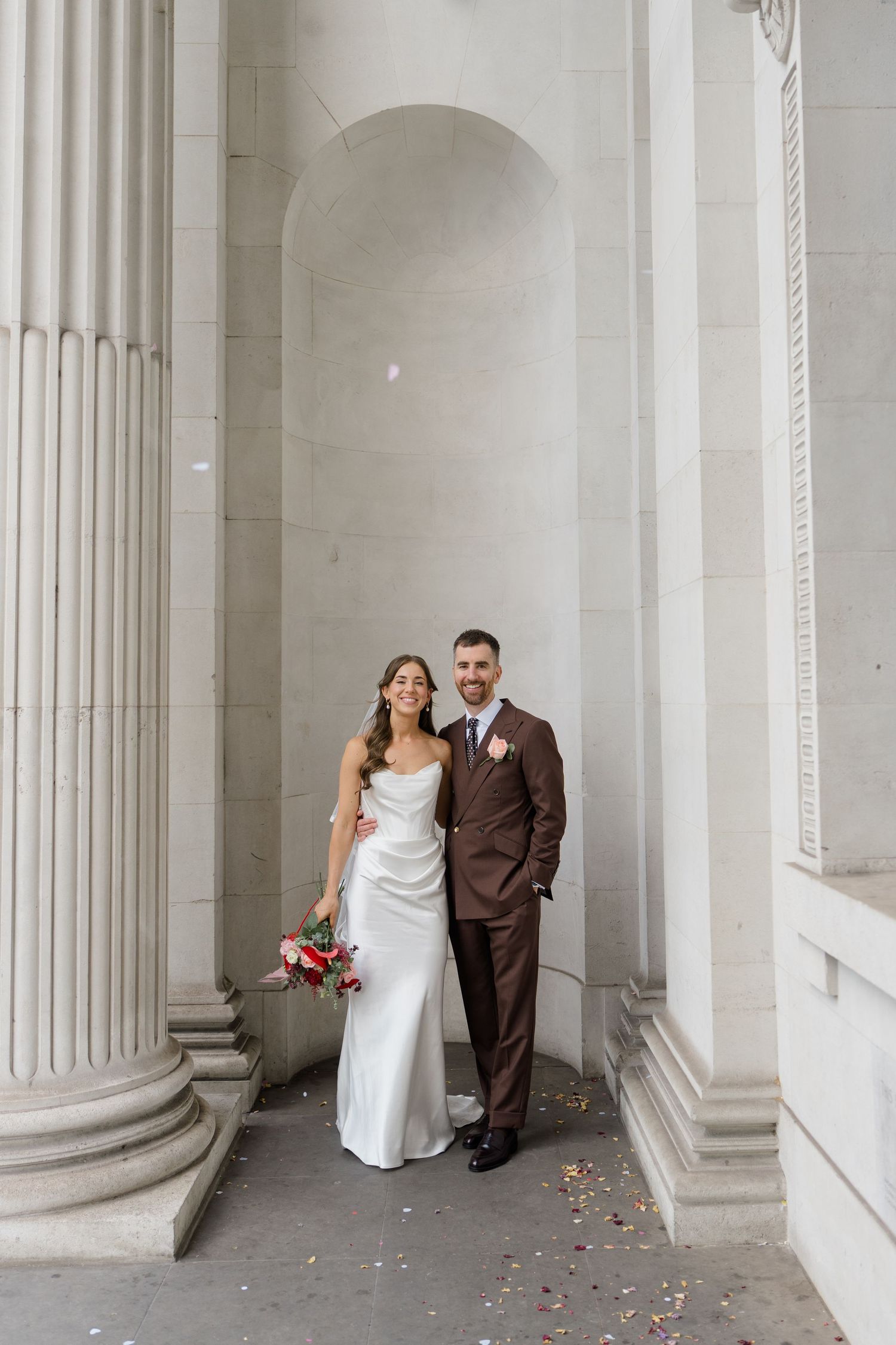 Bride and groom posing between grand stone columns, bride in white satin gown holding red bouquet, groom in brown suit.