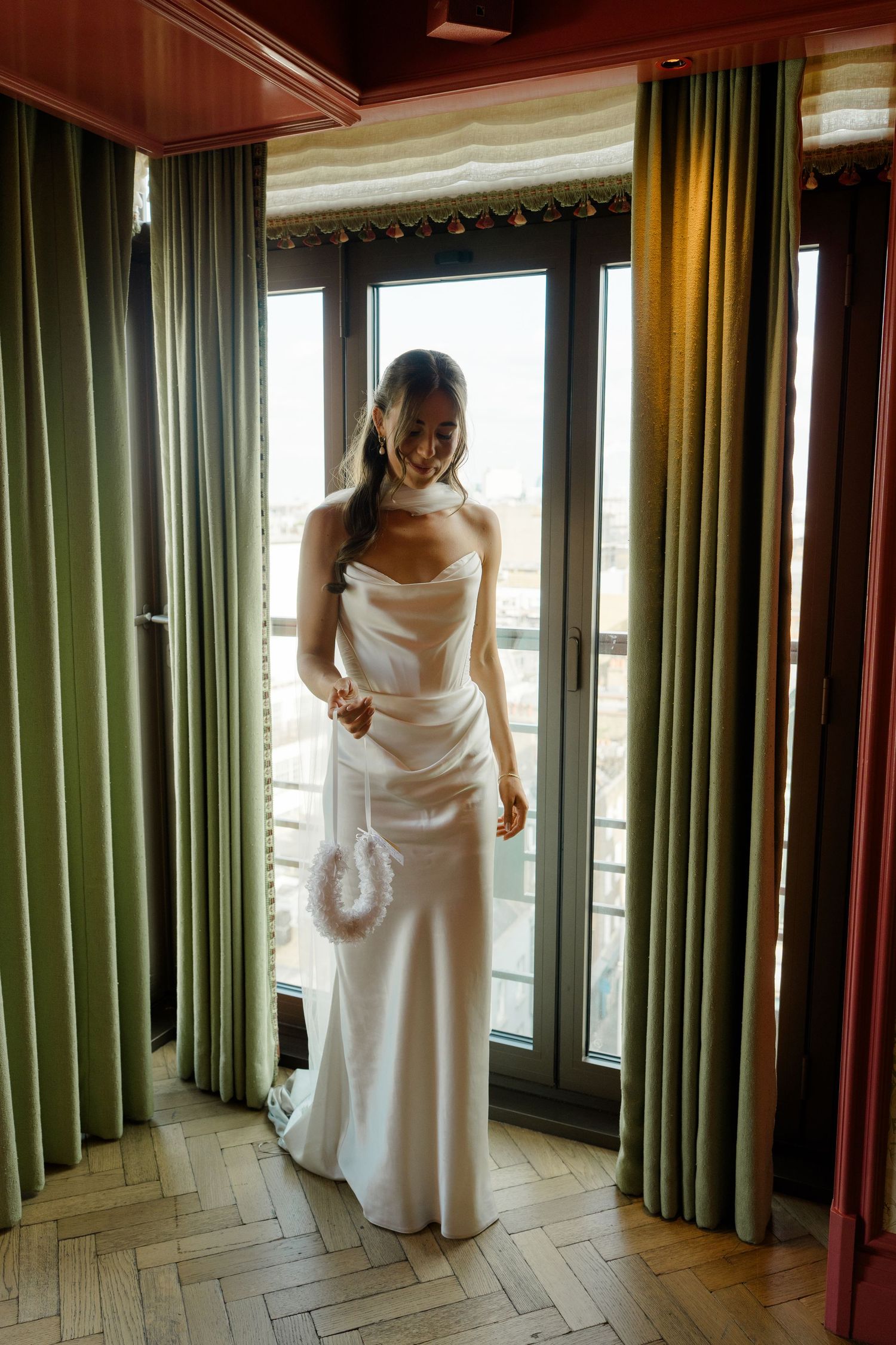 Bride in strapless white satin wedding gown stands by green curtained window holding feather accessory.
